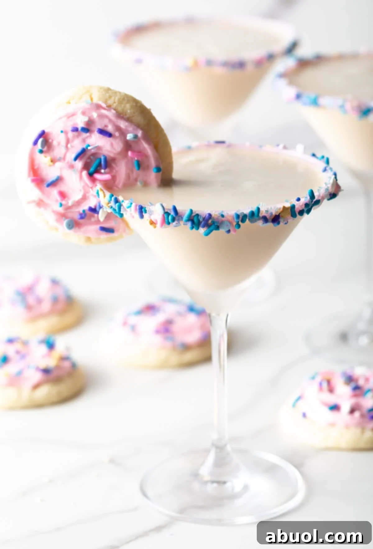 Finished sugar cookie martini surrounded by iced sugar cookies with a sugar cookie on the rim, showcasing the final presentation and deliciousness of the cocktail.
