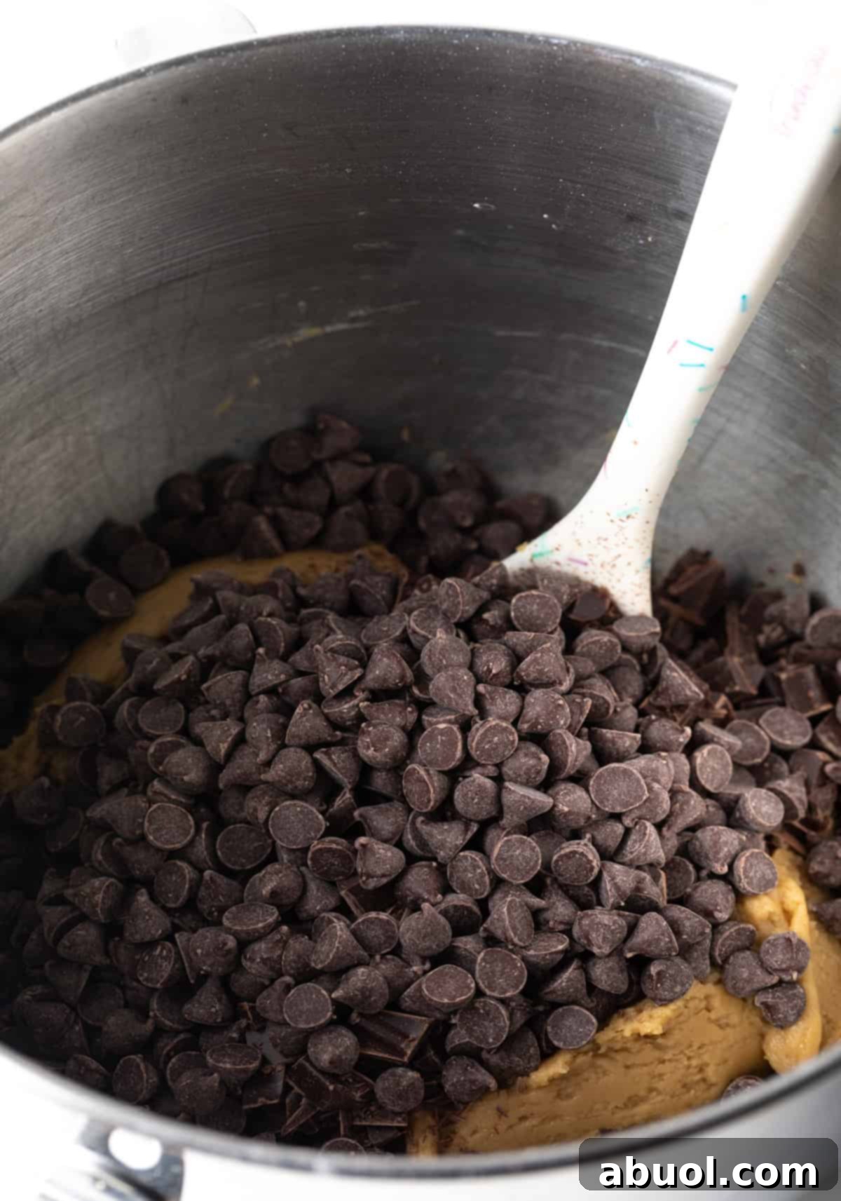 Chocolate chips being added to brown sugar cookie dough in a metal mixing bowl.