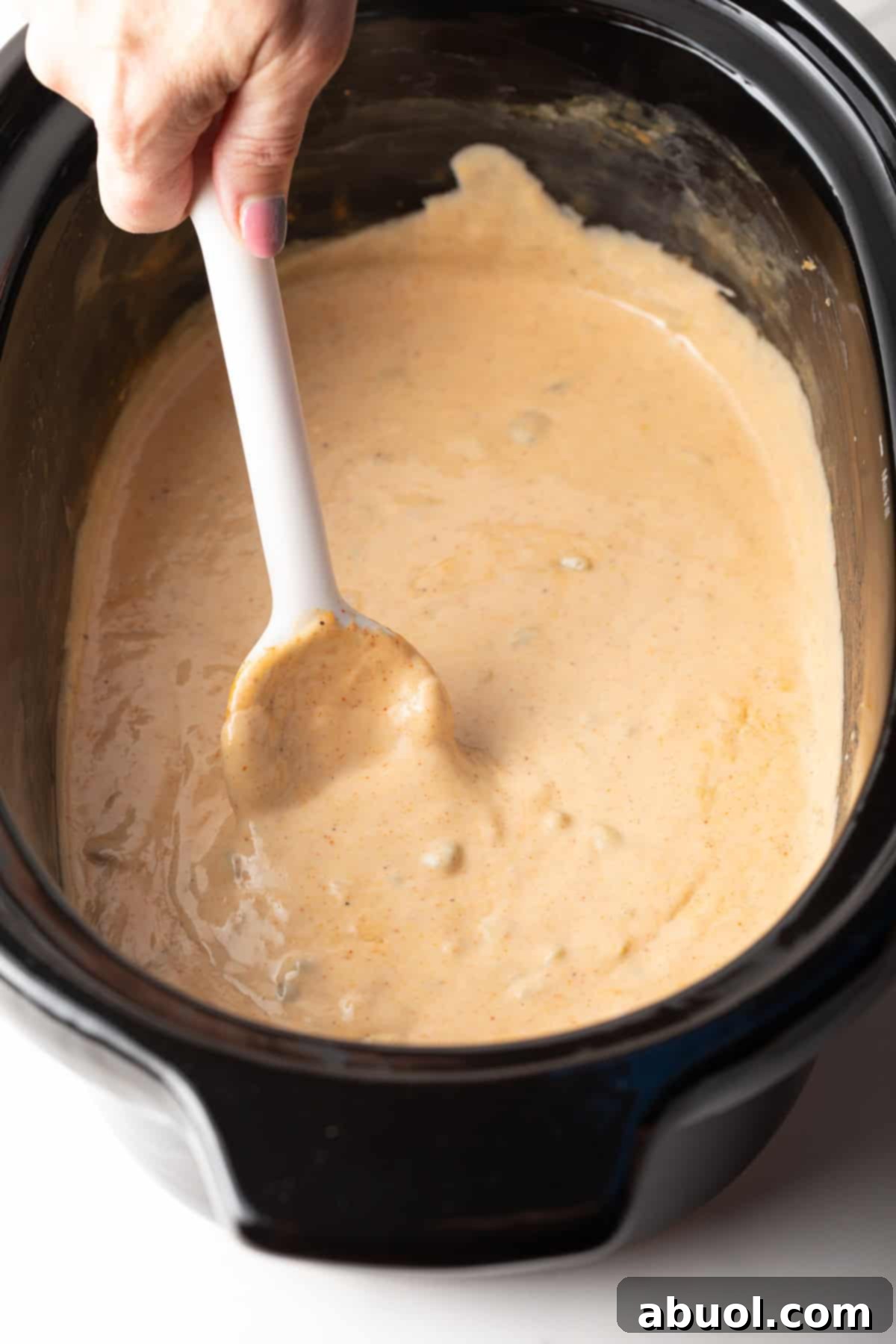 A white spoon stirring creamy slow cooker queso dip in a black crock pot.