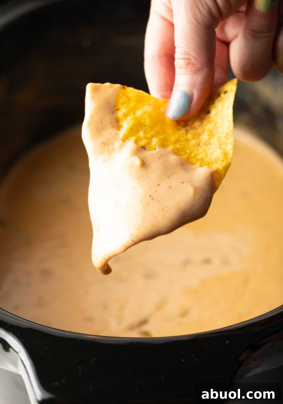 A hand holding a tortilla chip dipped in crock pot queso recipe over the crock pot.