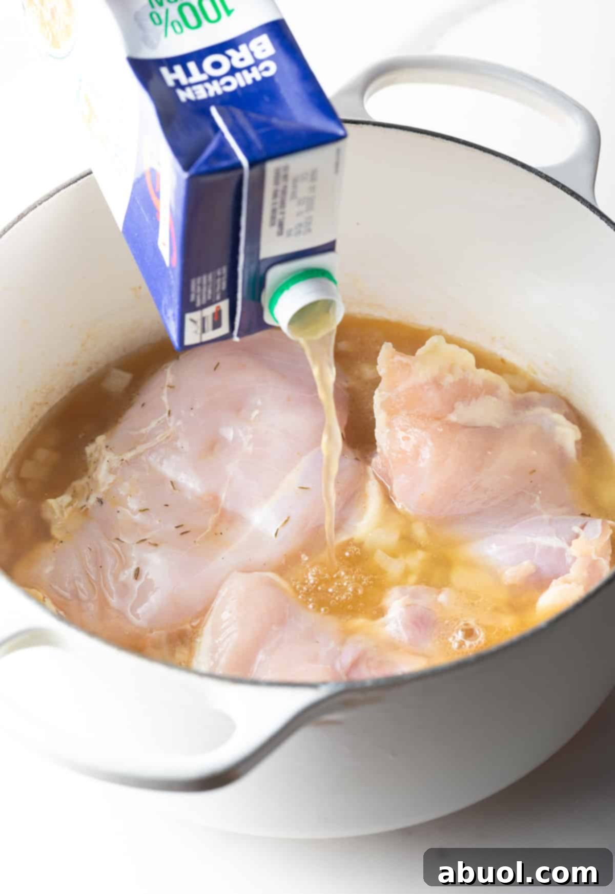 Pouring rich chicken broth from a carton into a soup pot containing raw chicken breasts.