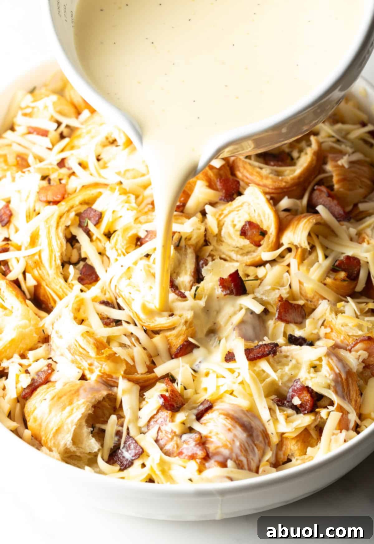 A cream and egg mixture is being added to a baking dish filled with unbaked croissant casserole.