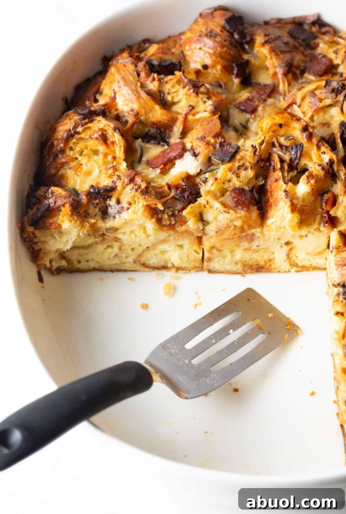 A metal spatula is serving slices of croissant casserole from a white baking dish.
