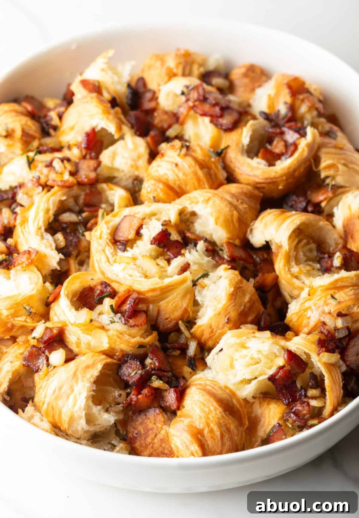 A bacon and vegetable mixture is being added to torn croissants in a white baking dish.
