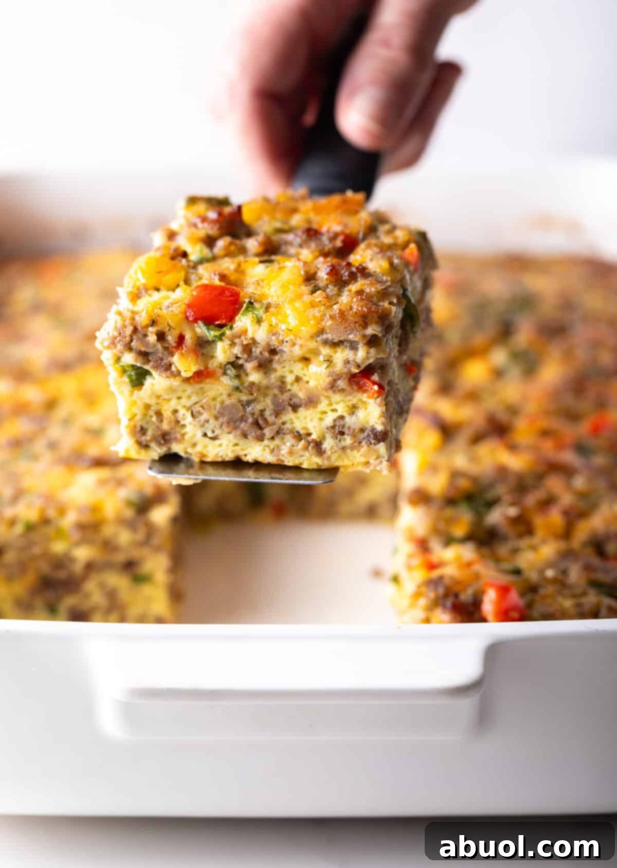 Sausage and vegetable egg casserole in a pan with a hand holding a spatula above it. Square portions have been cut.