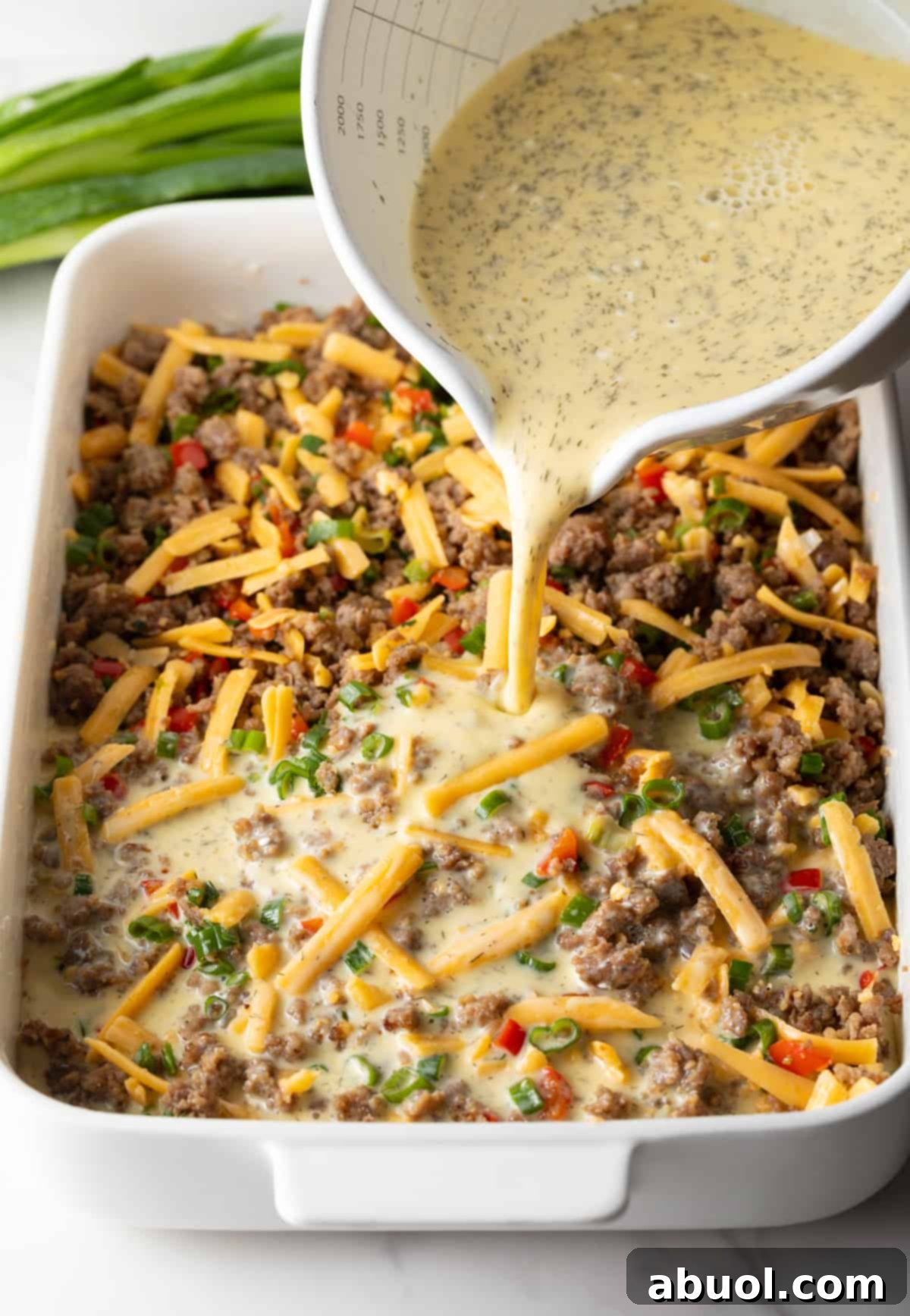 Pouring egg mixture over shredded cheese and sausage in a white rectangle baking dish
