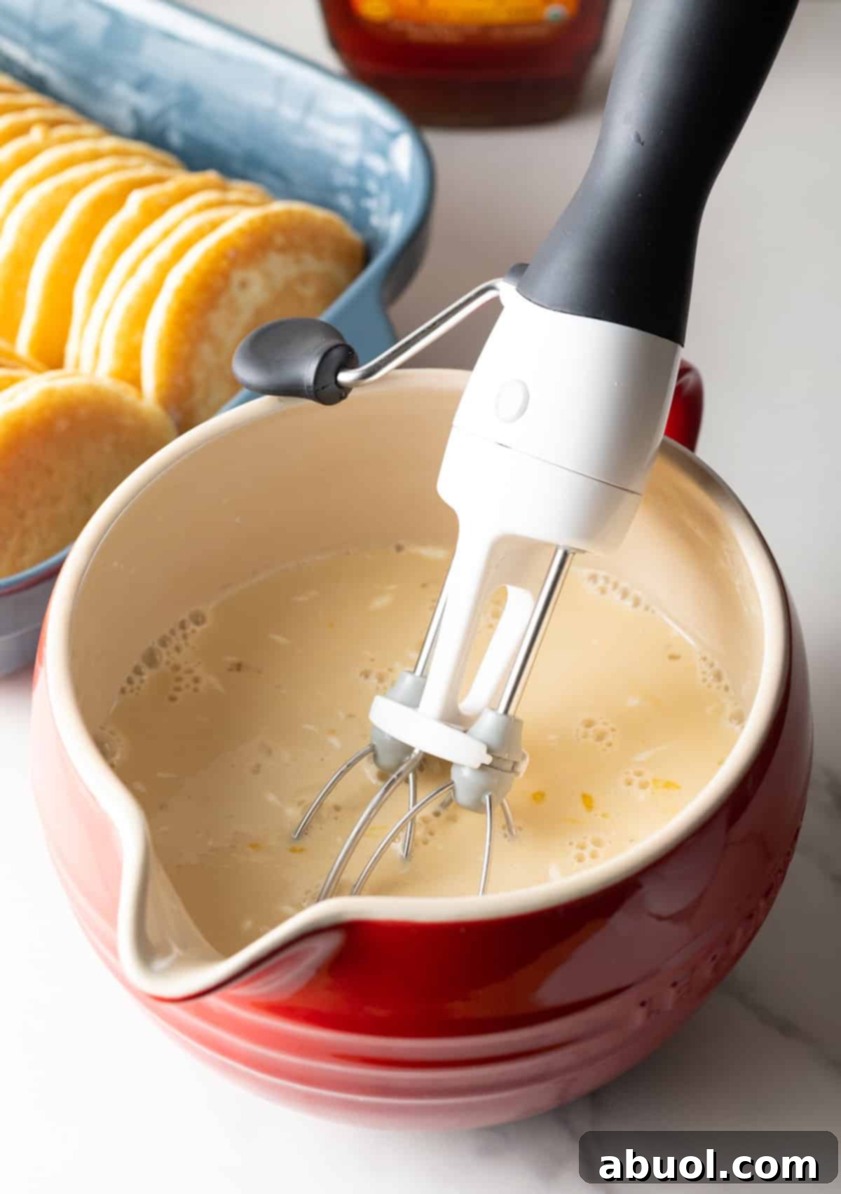 A whisk mixing a cream and egg mixture in a red bowl.