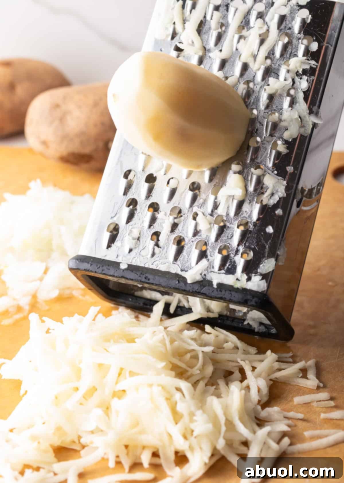 A peeled potato is being shredded with a box grater onto a wooden cutting board.