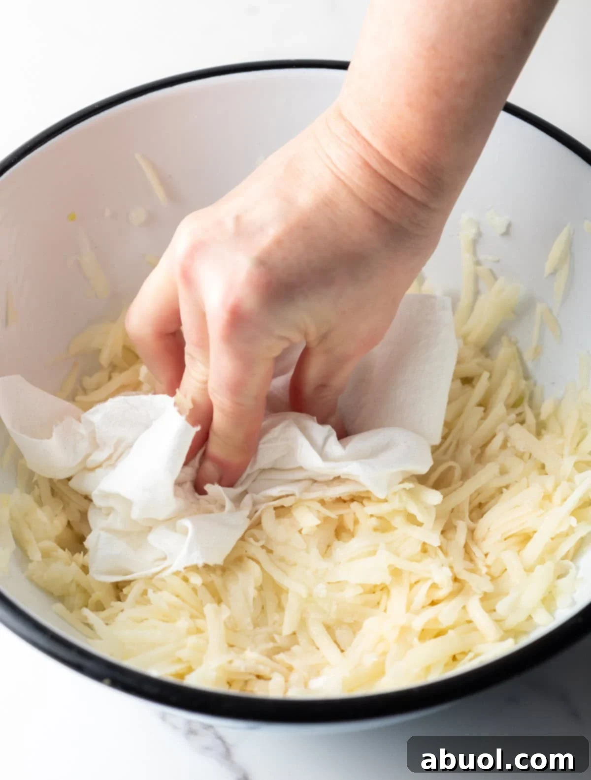 A hand holding a paper towel that is being used to remove moisture from shredded potatoes for a homemade hash brown recipe.