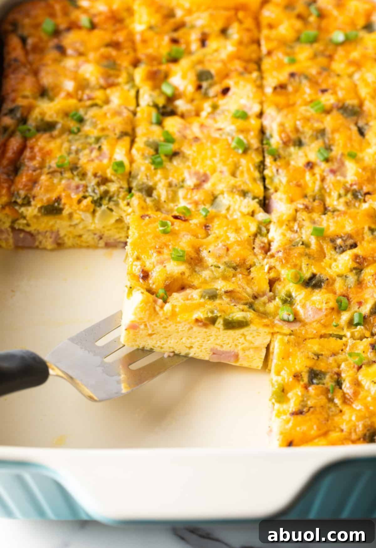 A slice of a western omelette casserole is being scooped by a metal spatula over a white baking tray filled with a Denver omelette casserole.