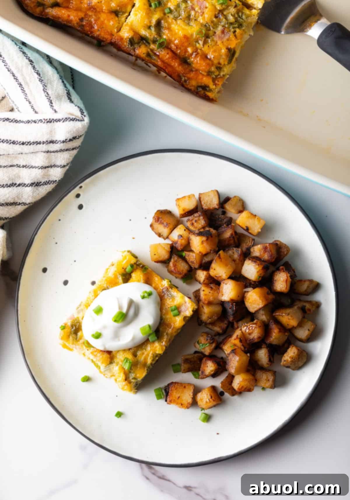 An overhead shot of a slice of baked Denver omelet casserole topped with sour cream, next to a serving of home fries on a white plate.