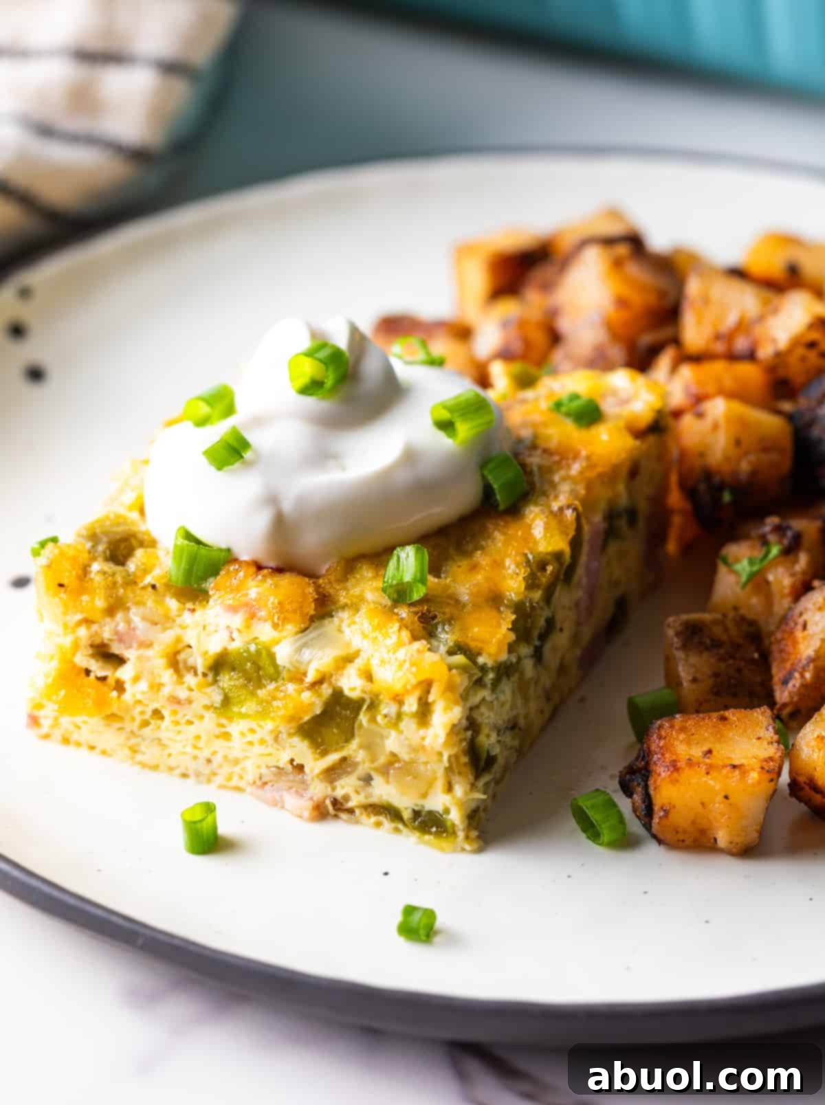 A slice of Denver baked omelet casserole topped with sour cream, next to a serving of home fries on a white plate.