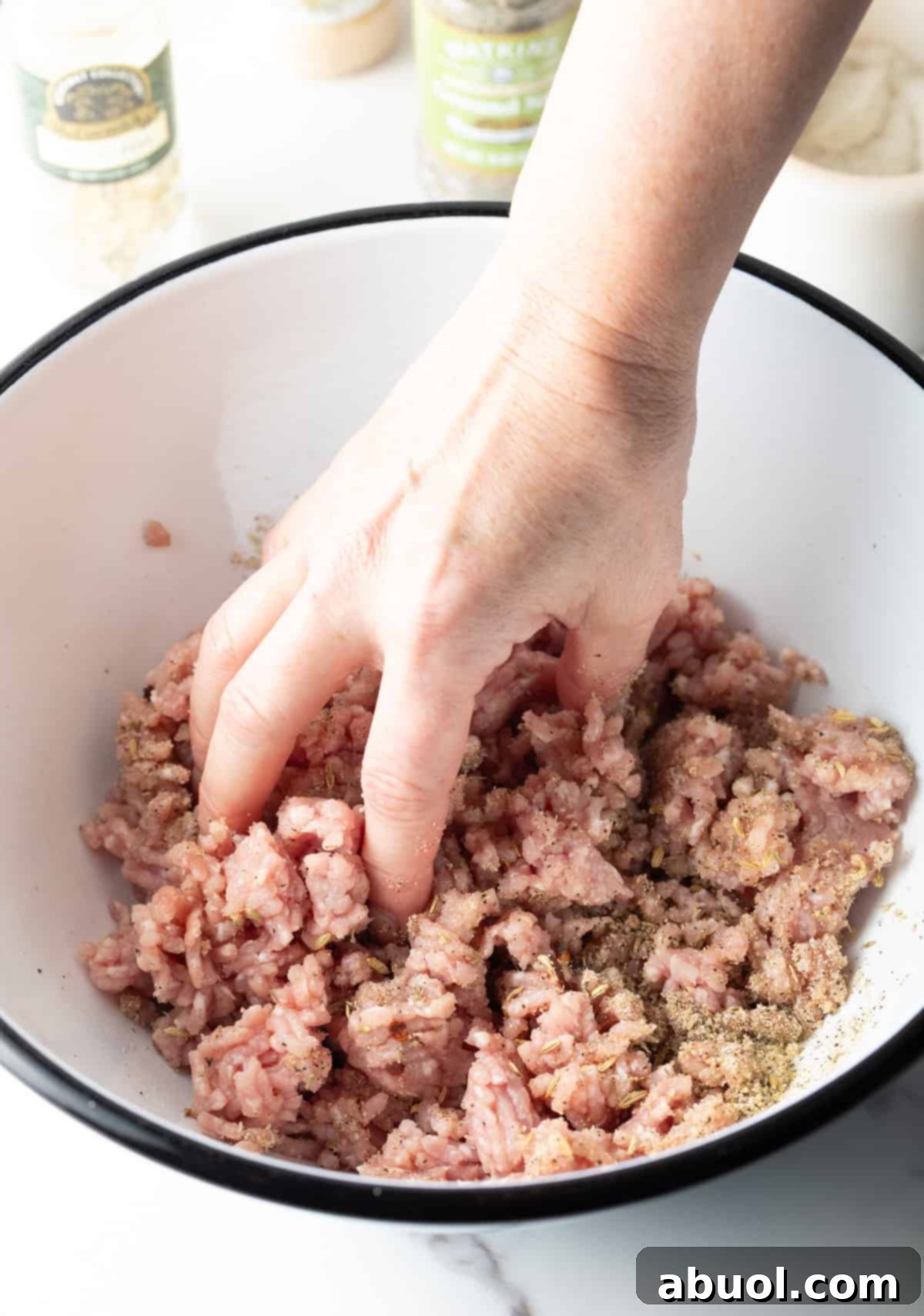 A hand gently mixing ground pork with a spice blend in a white bowl to make homemade sausage.