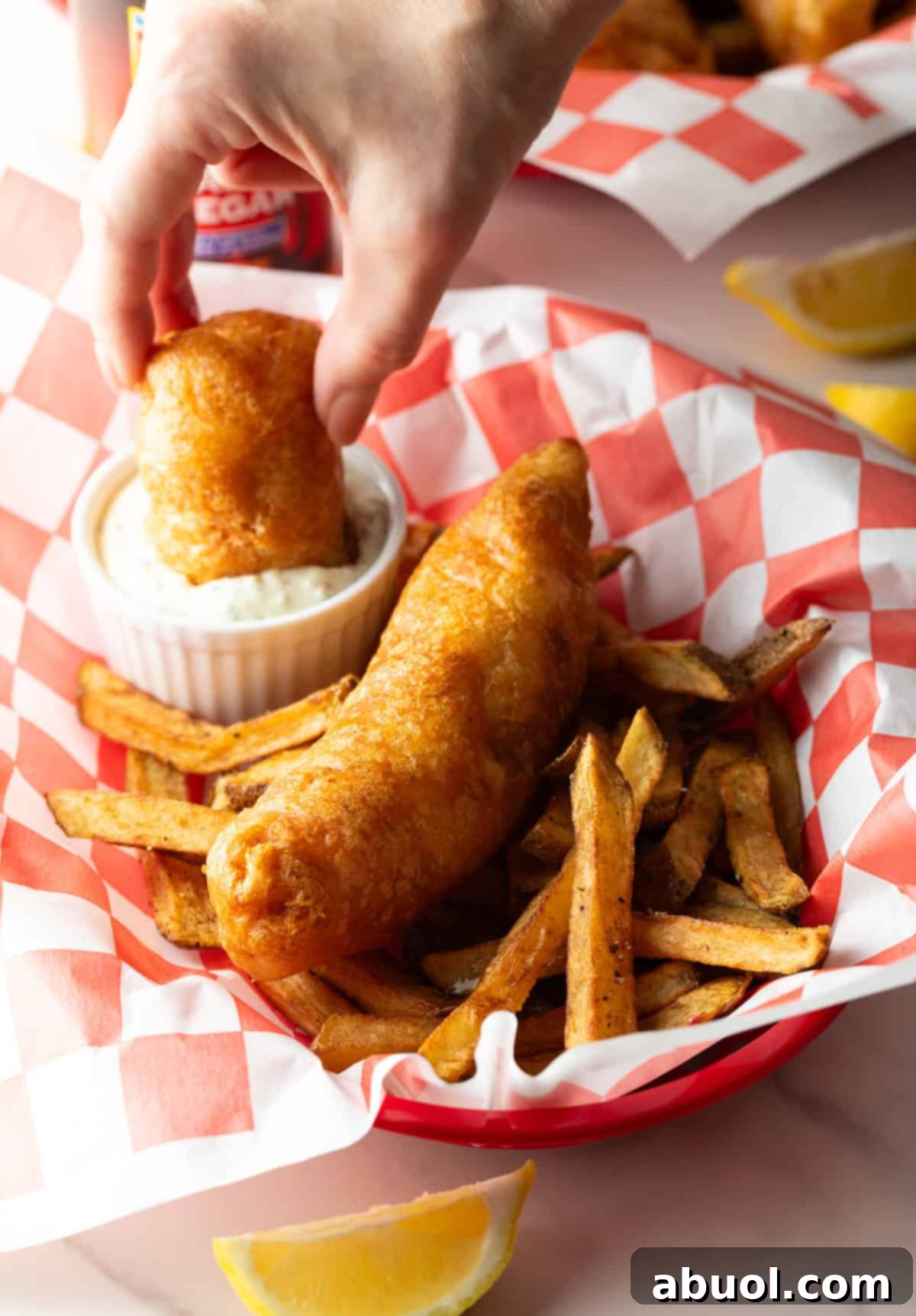 A hand dunking a piece of crispy fried fish into a small white ramekin filled with tartar sauce.