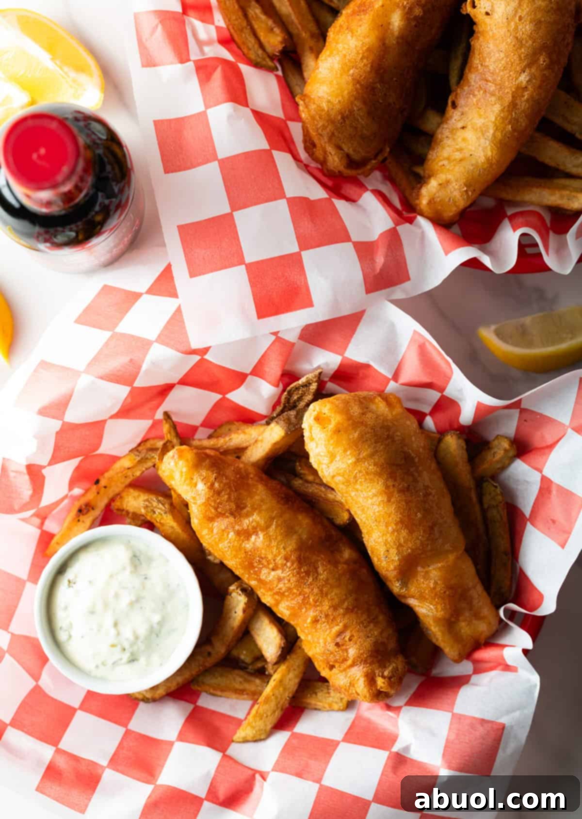 An overhead view of a paper-lined basket filled with beer-battered fried fish and french fries, with a side of sauce.