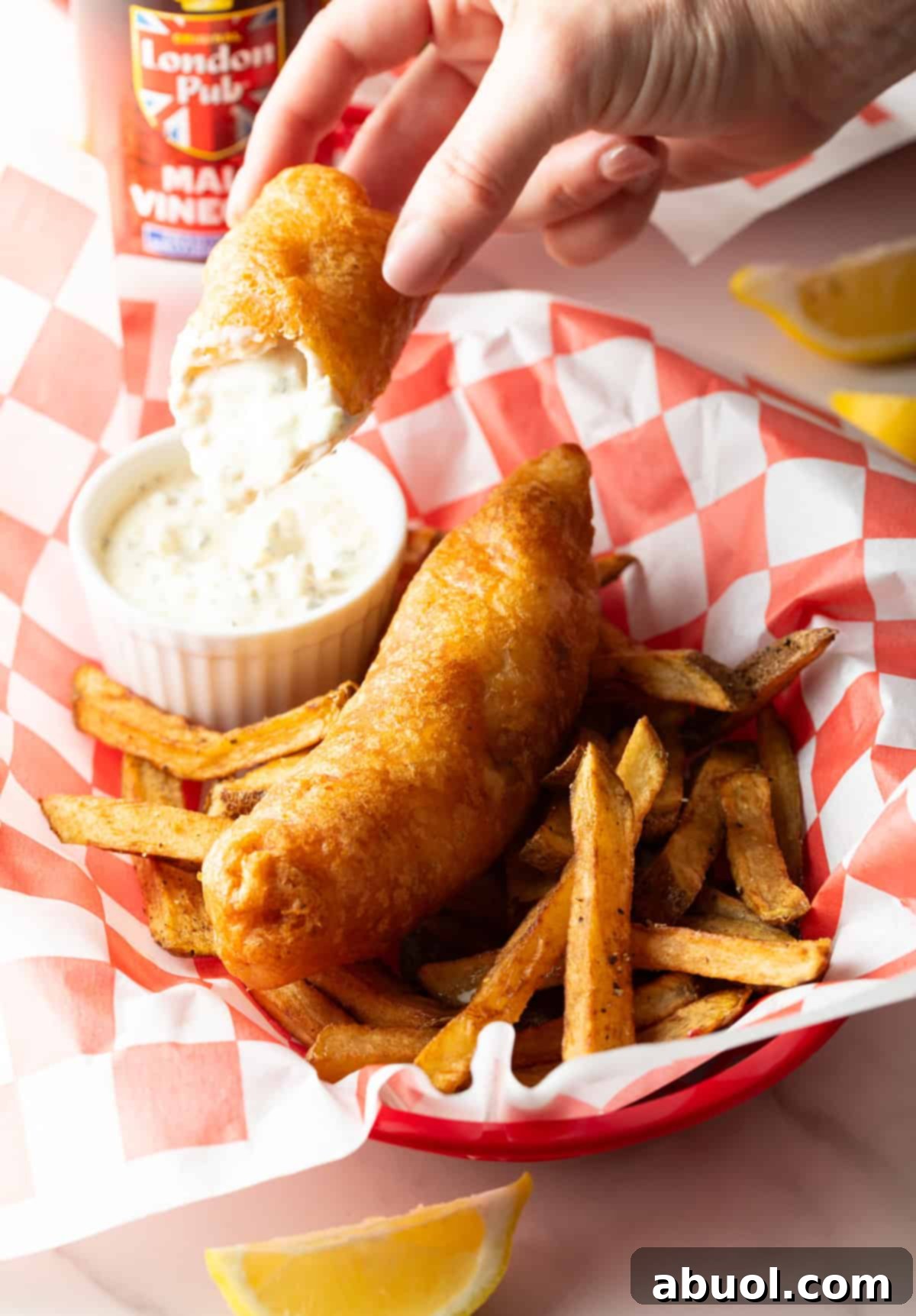 A beautiful serving of fish and chips in a basket, with one piece of fish being dipped into a side of sauce.
