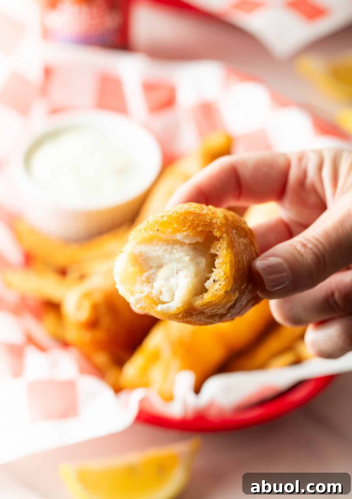 A hand holding a piece of golden beer-battered fried fish with a bite taken out, revealing the flaky white fish inside.
