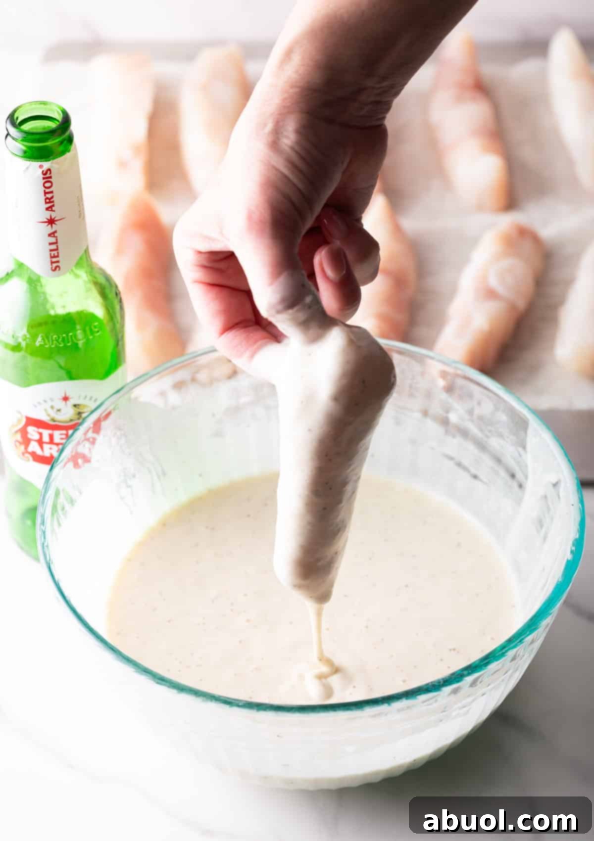 A hand dipping a piece of raw fish fillet into the prepared beer batter.