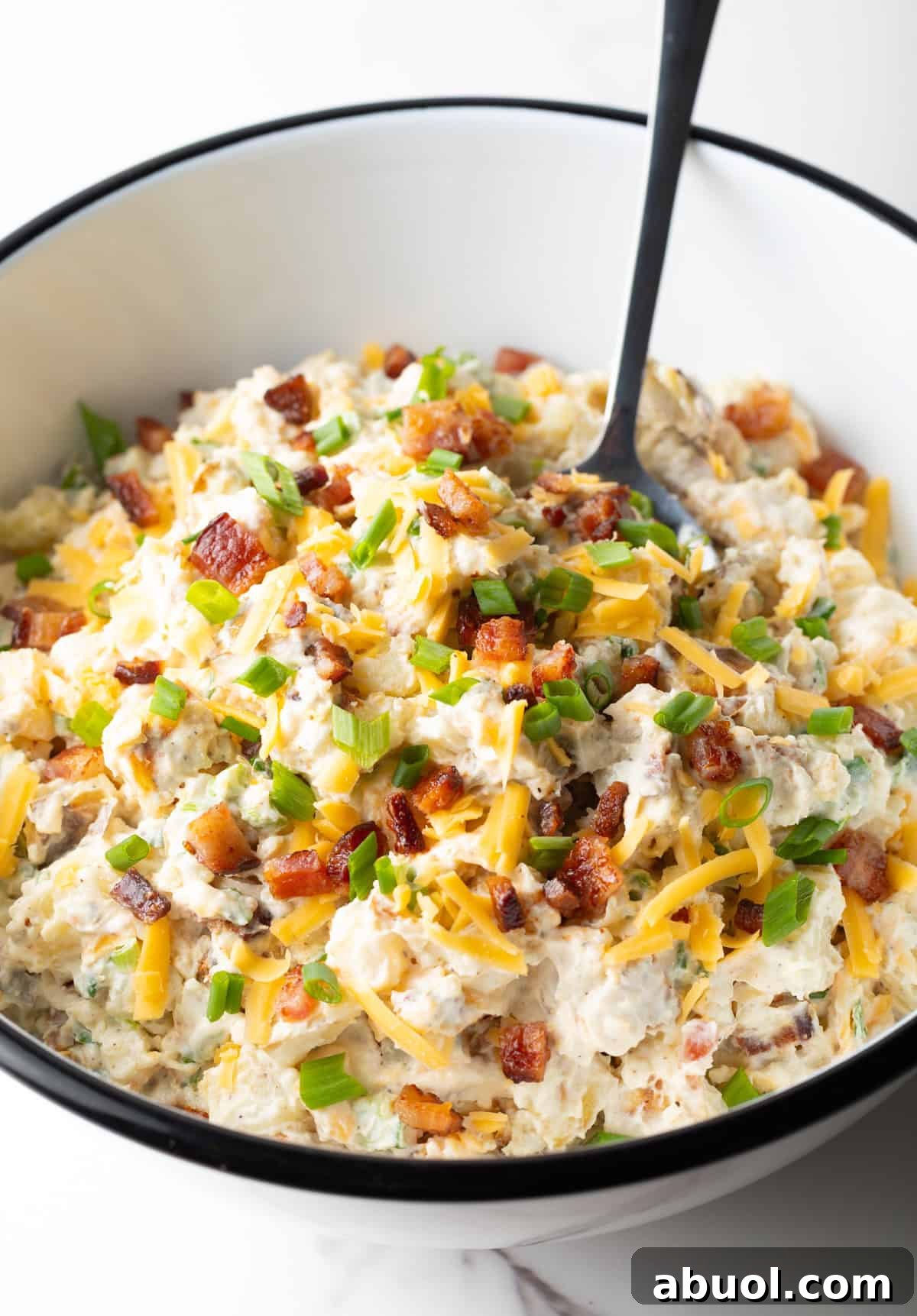 A loaded baked potato salad recipe topped with bacon, cheese, and chives, sitting in white bowl with a spoon.