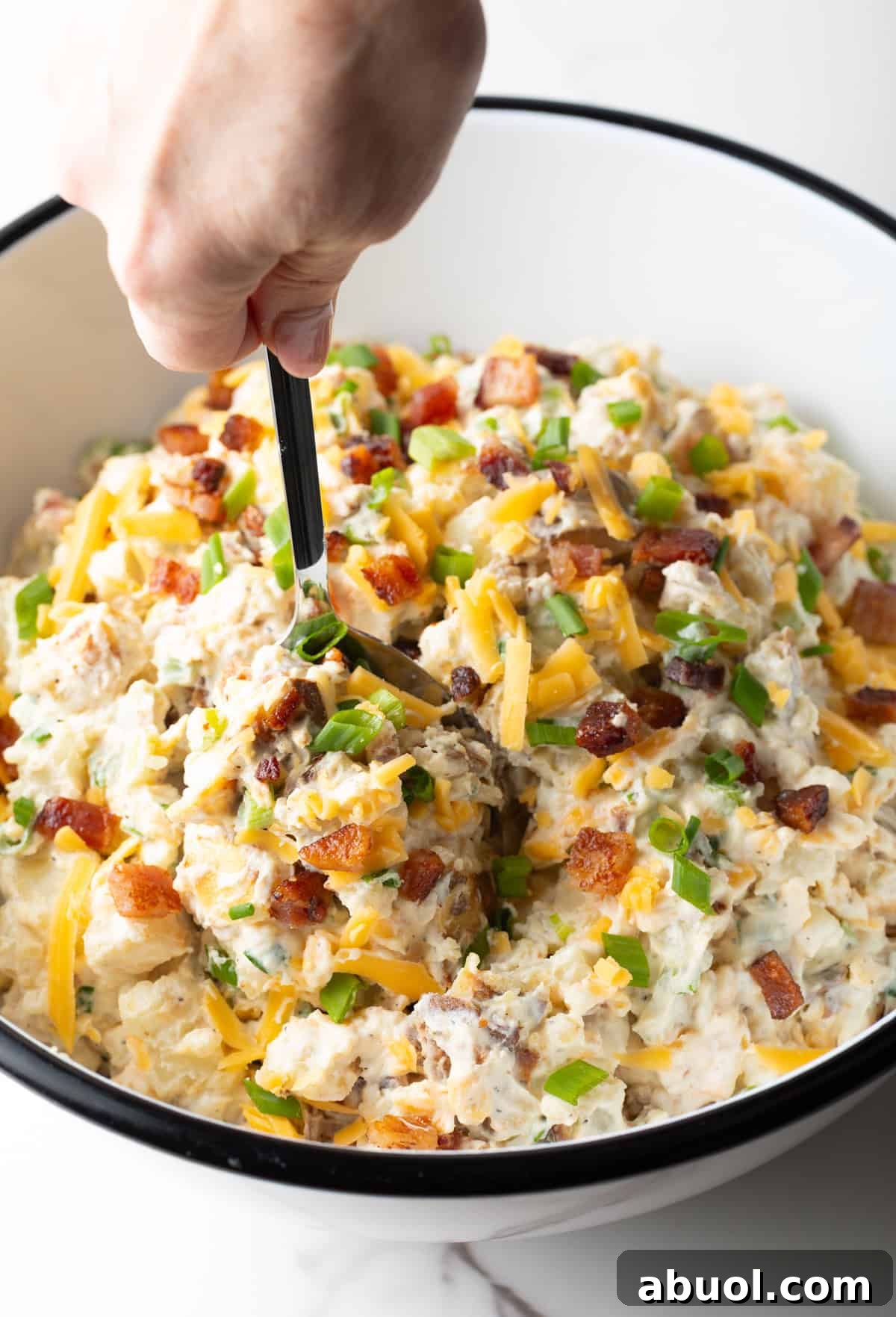 A hand holding a spoon is scooping a serving of baked potato salad from a bowl.