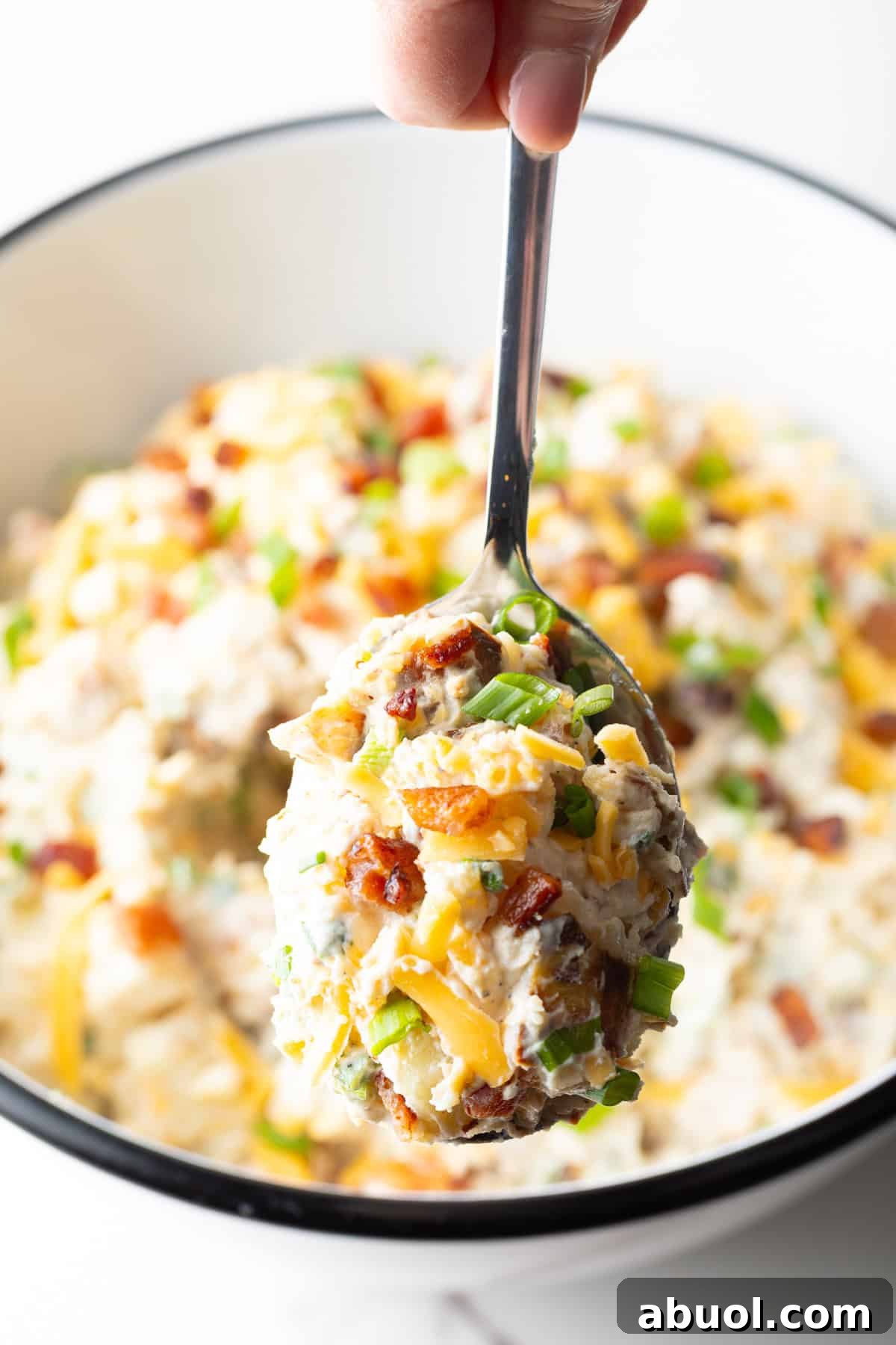 A spoon holding a scoop of loaded baked potato salad over a bowl filled with the salad.