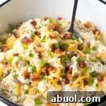 A loaded baked potato salad recipe topped with bacon, cheese, and chives, sitting in white bowl with a spoon.