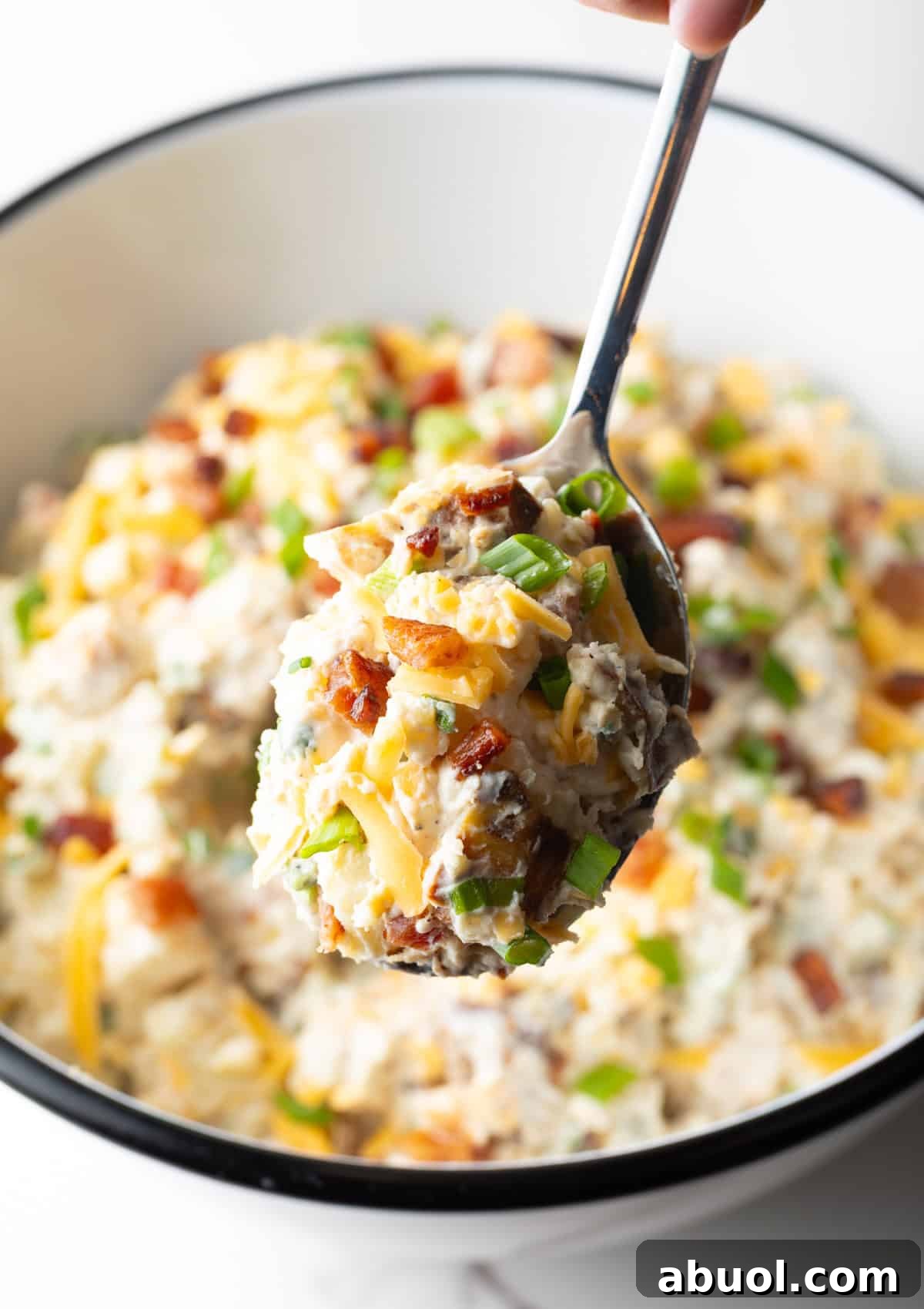 A spoon holding a scoop of baked potato salad over a bowl filled with the salad.