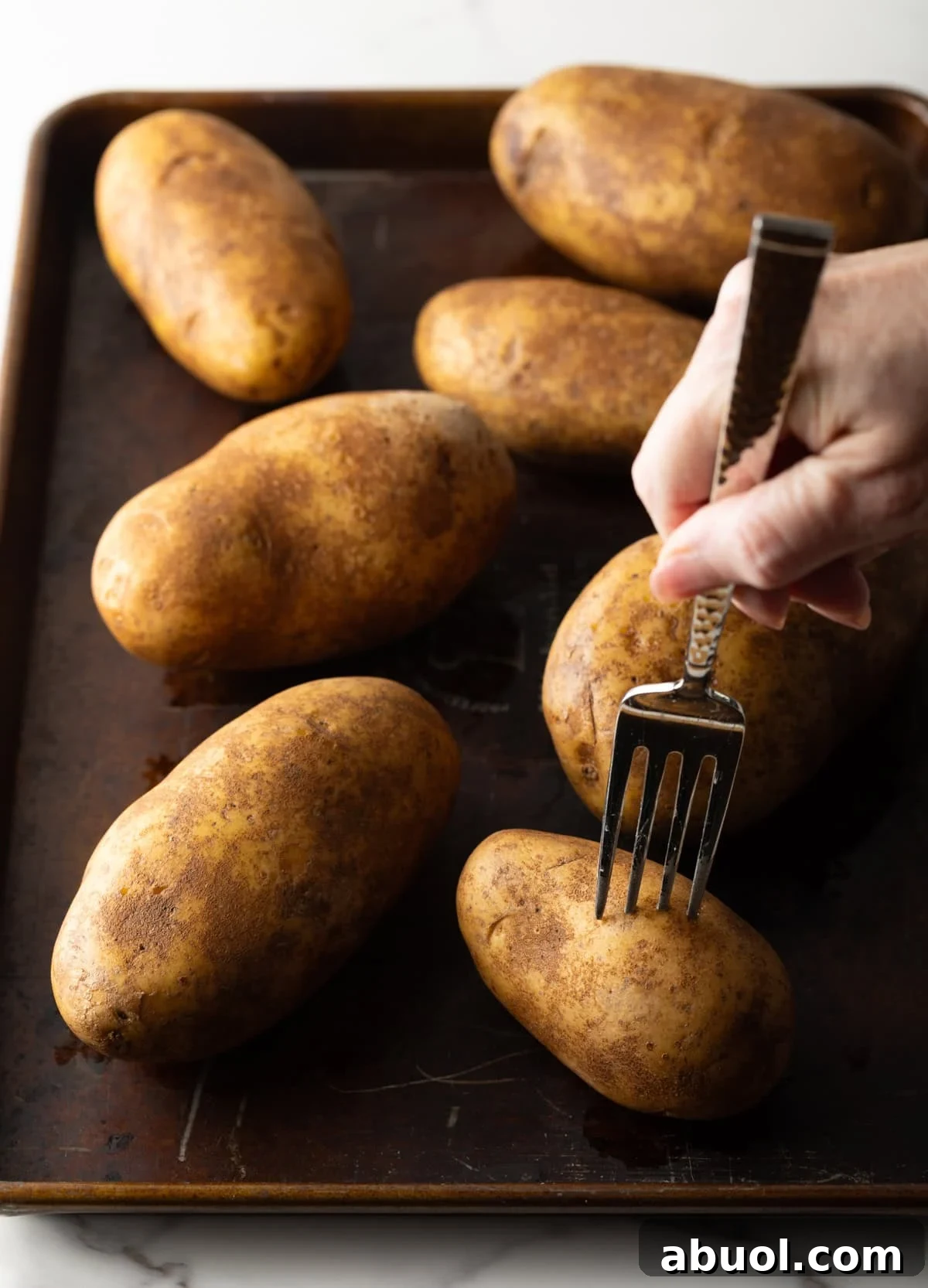 A fork is poking whole russet potatoes on a baking sheet.