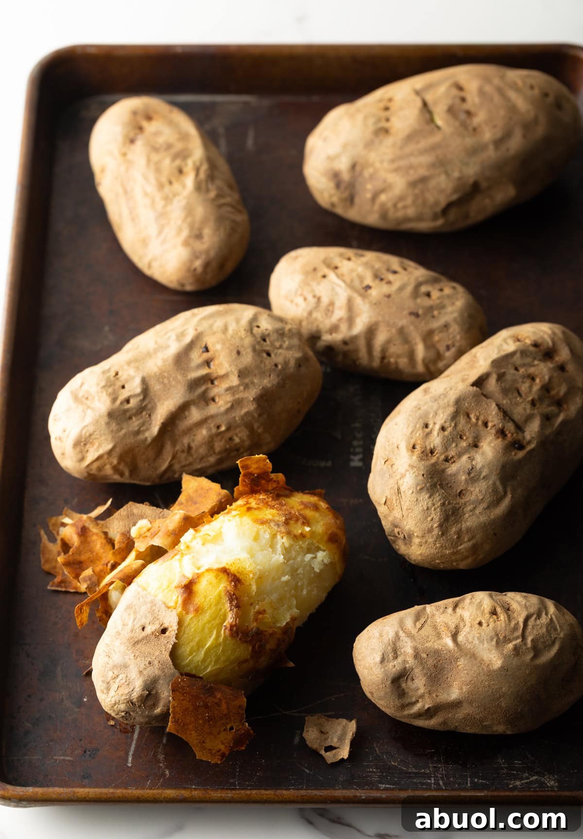 Baked potatoes on a baking sheet, one of which is mostly peeled.