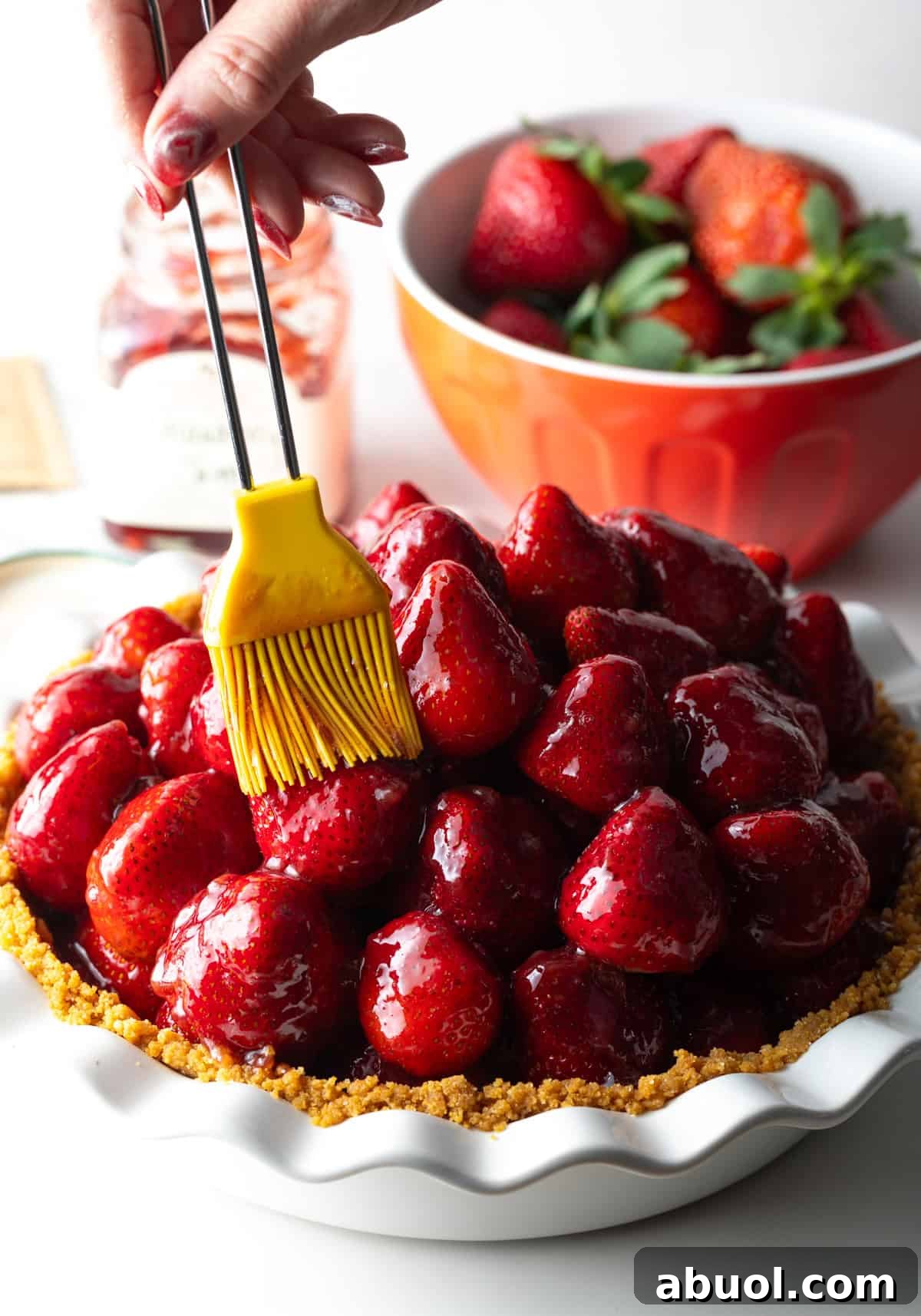 A pastry brush is brushing a strawberry jam mixture over a fresh strawberry pie recipe.