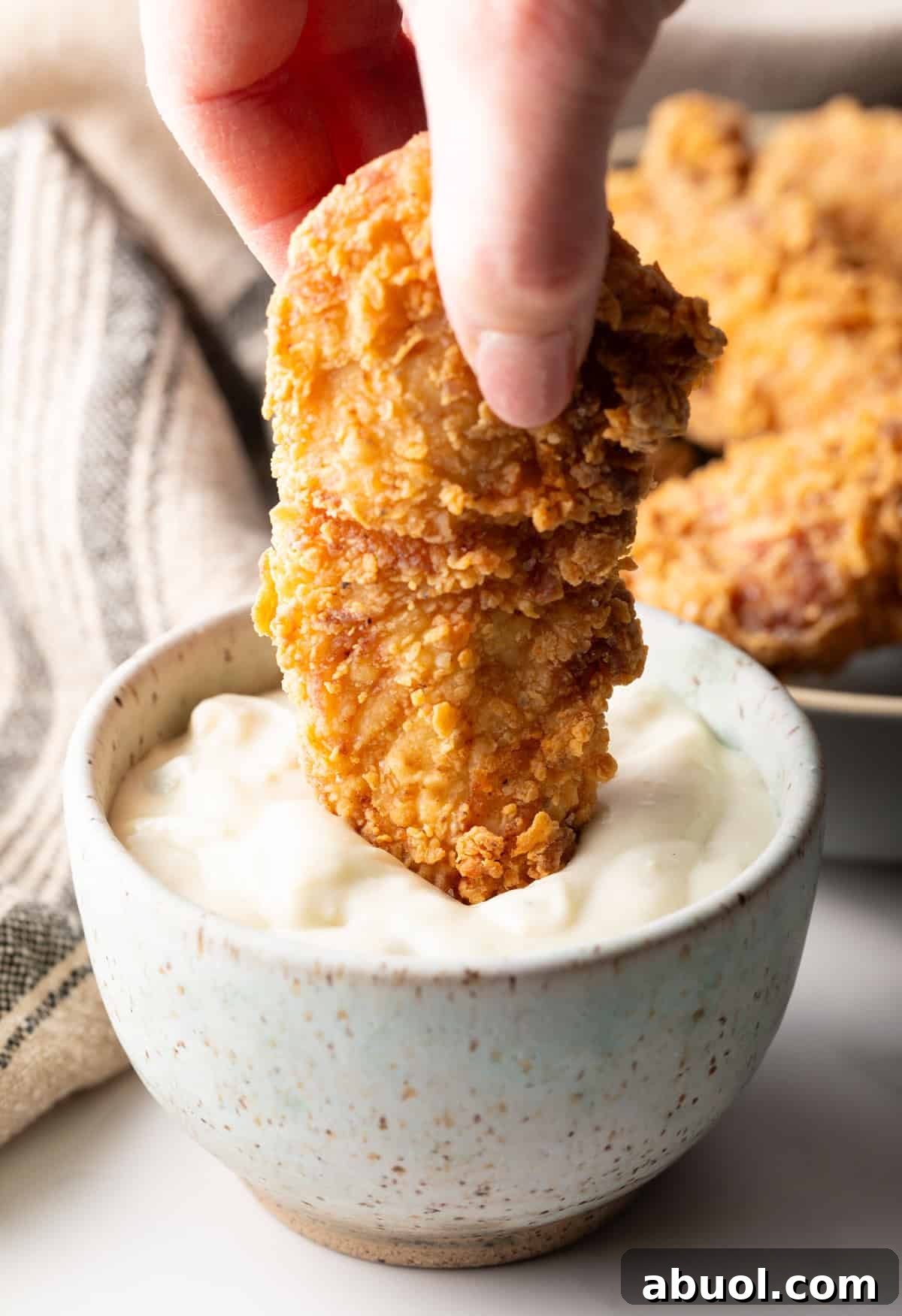 A homemade fried chicken tender is being dipped into a ramekin of white sauce.