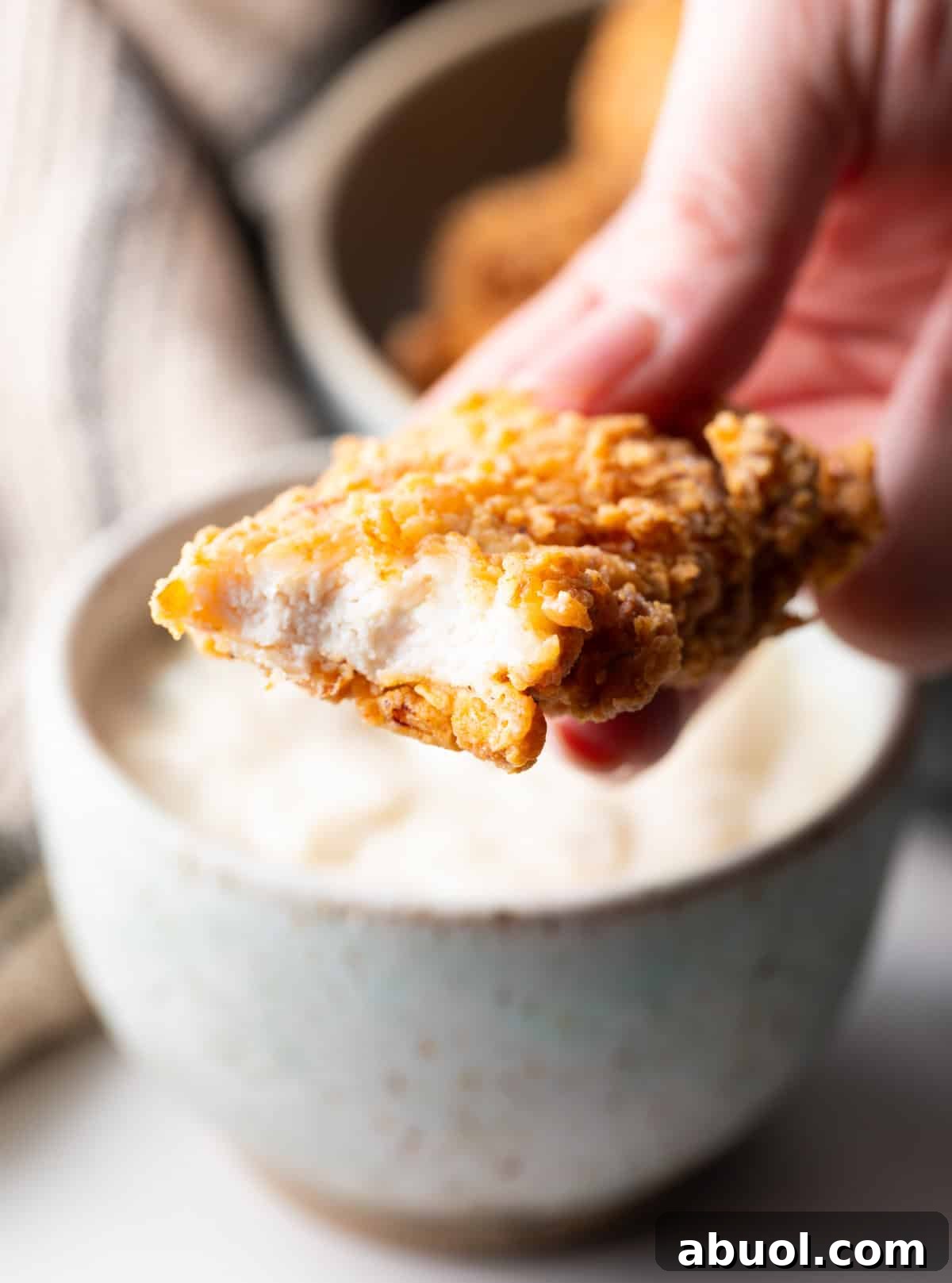 A hand holding a deep fried chicken tender with a bite taken out.