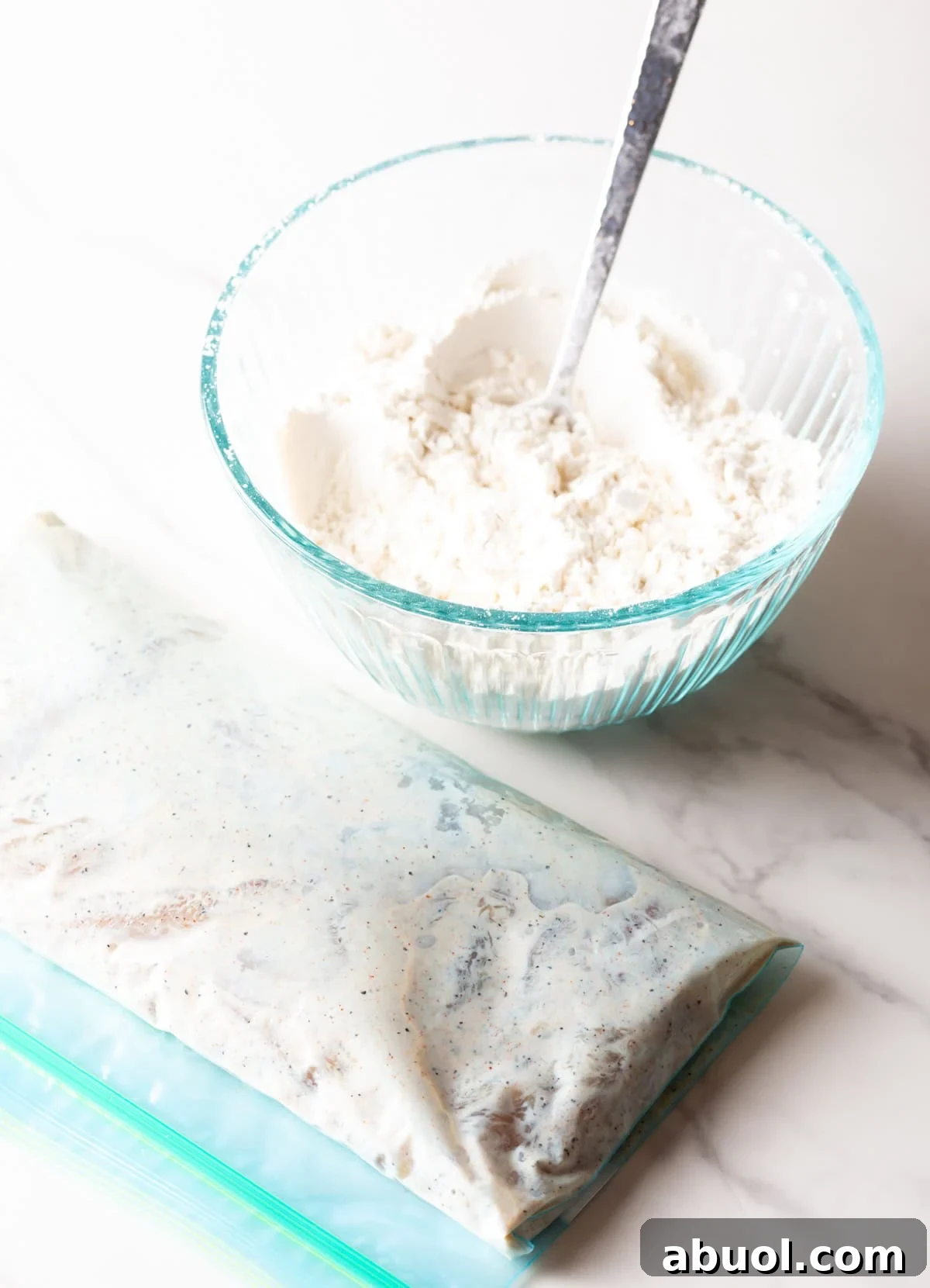 A glass bowl filled with a flour mixture sitting next to a bag of raw chicken tenders marinating in a buttermilk mixture.