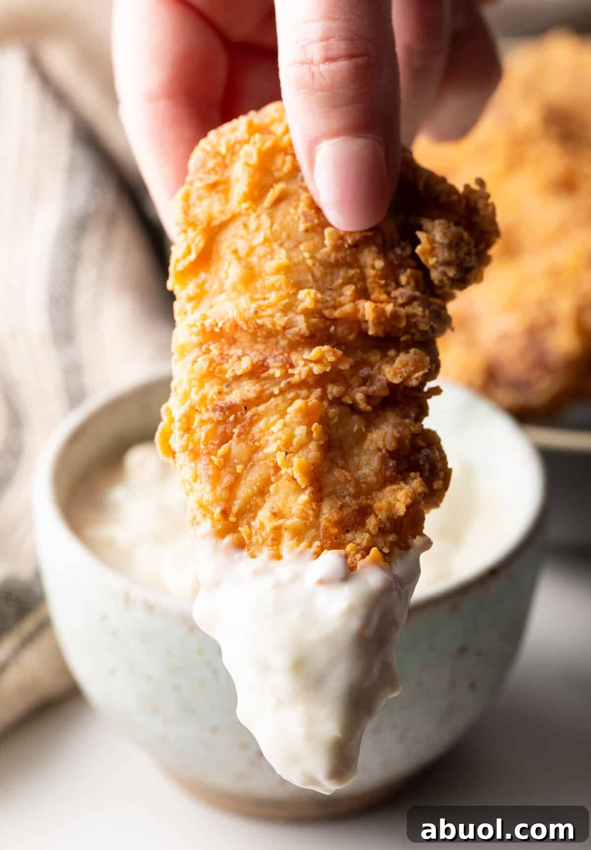 A homemade deep fried chicken tender is being dipped into a ramekin of white sauce, with the sauce dripping over the side of the ramekin.
