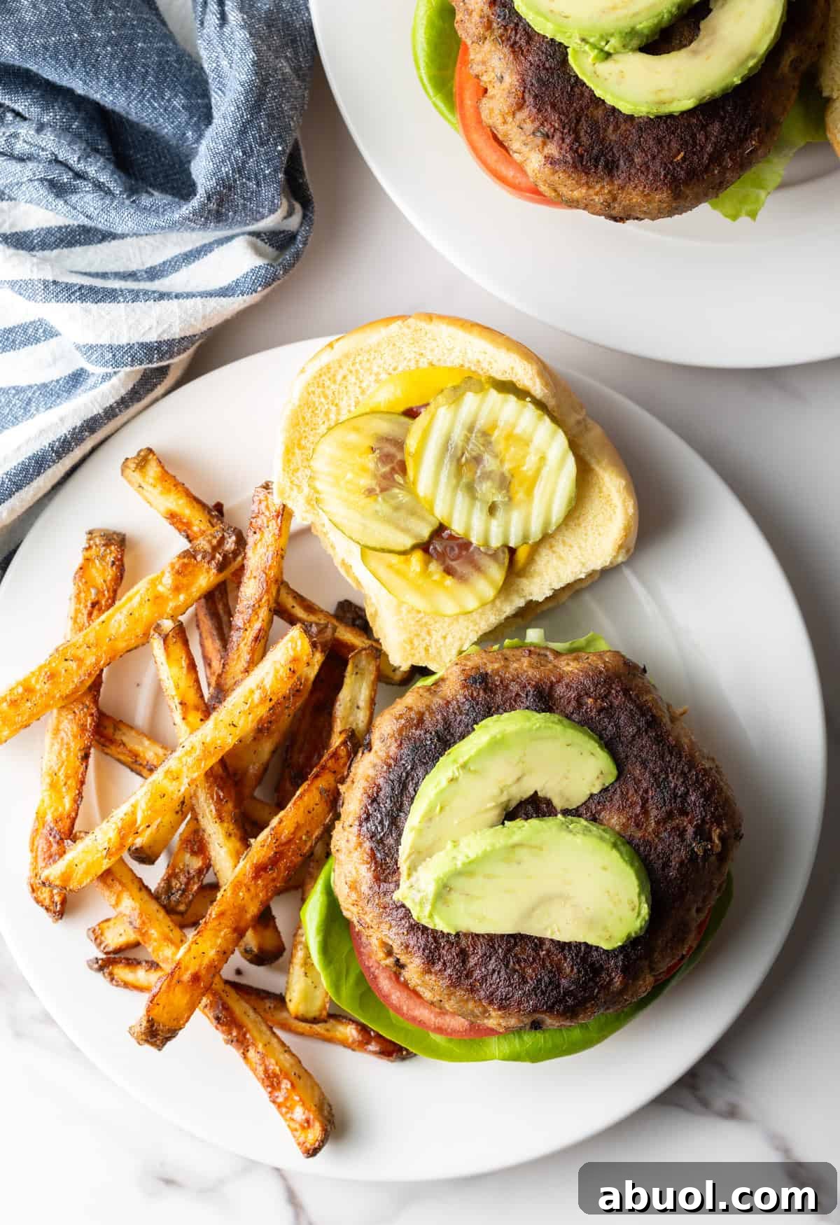 An overhead shot of a turkey burger with the top bun off, showing avocado and pickle slices, on a plate with fries.
