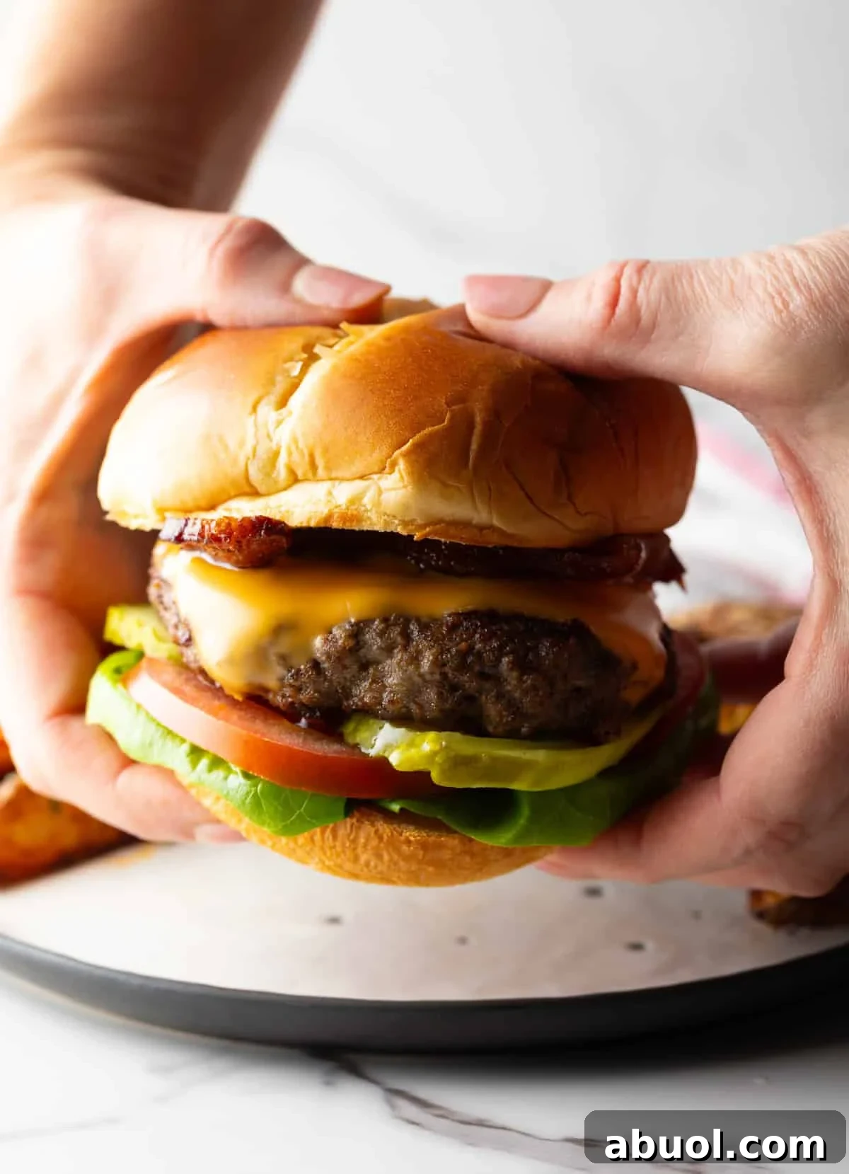 A hand is holding a fully assembled deer meat burger topped with cheese, lettuce, tomato, and pickles.
