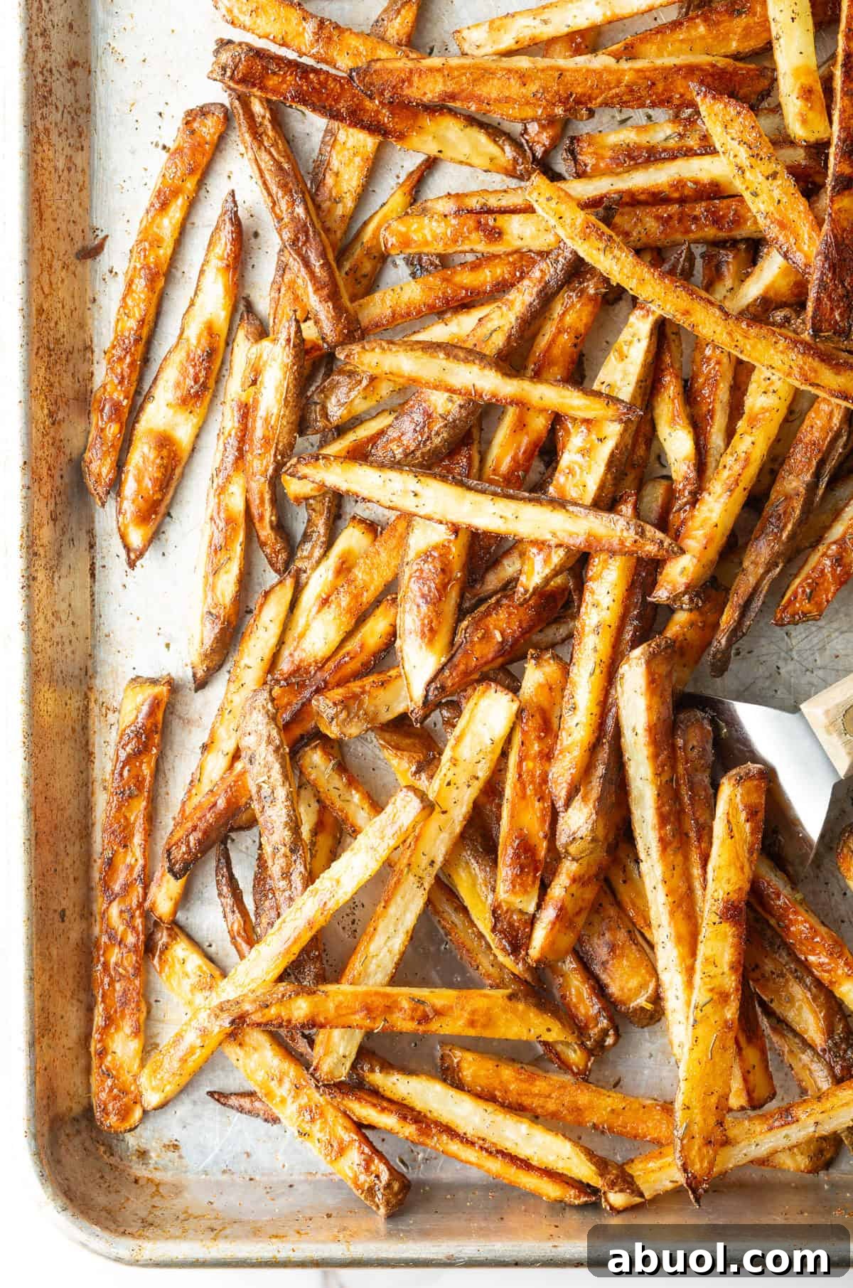 Crispy oven-baked French fries on a baking tray, being scooped by a metal spatula.