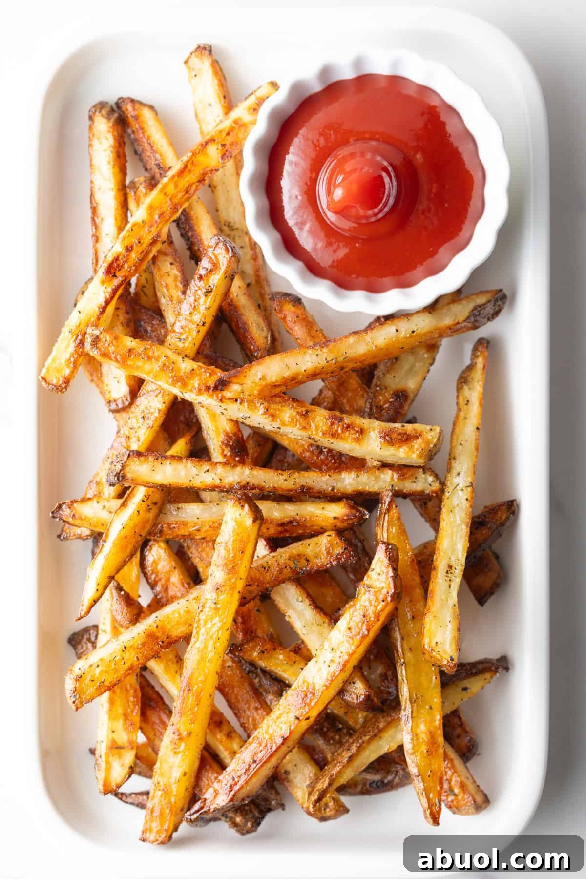 Homemade baked French fries from the oven sitting on a white plate with a ramekin of ketchup.