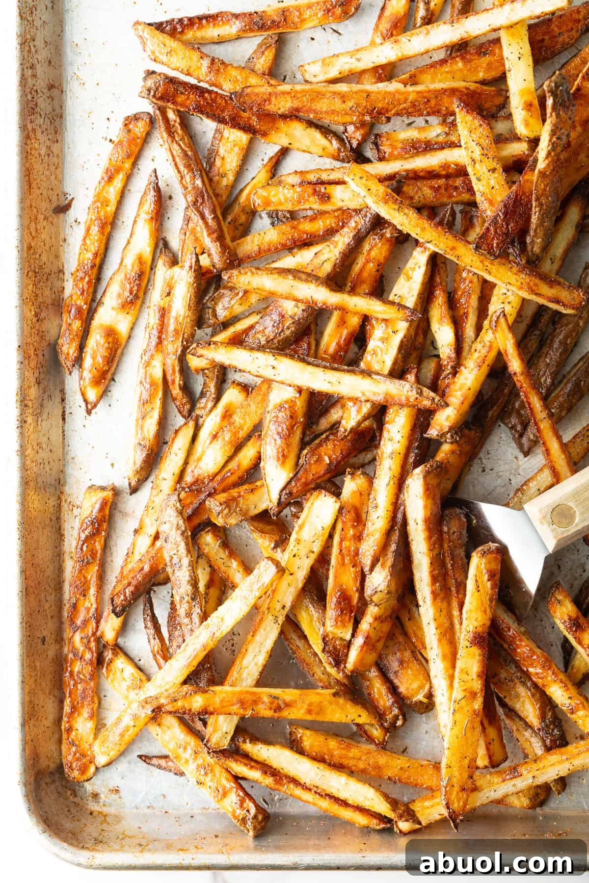 Oven baked French fries on a baking tray, being scooped by a metal spatula.
