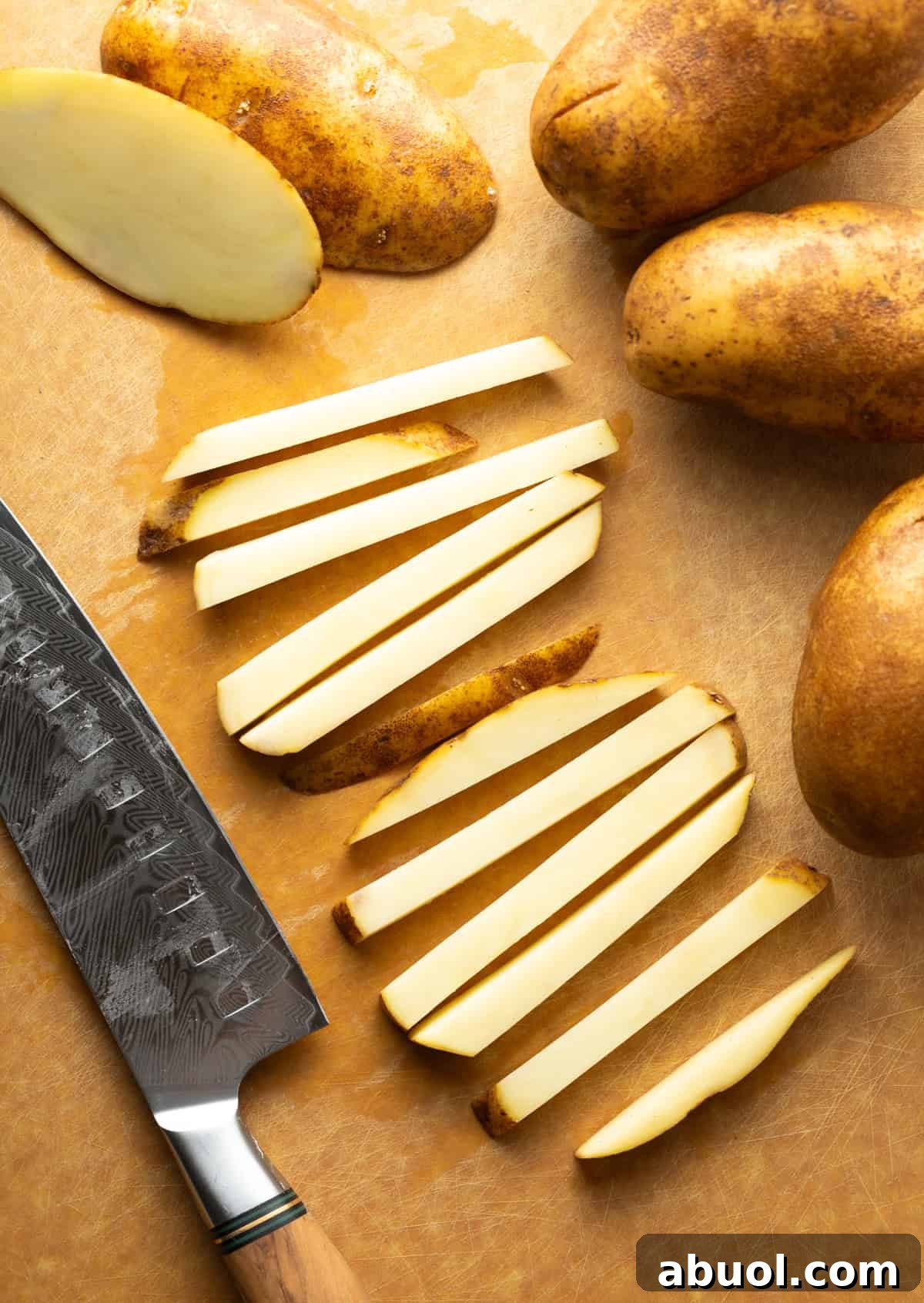 Potato slices on a wooden cutting board next to a knife, being prepared for a baked French fries recipe.