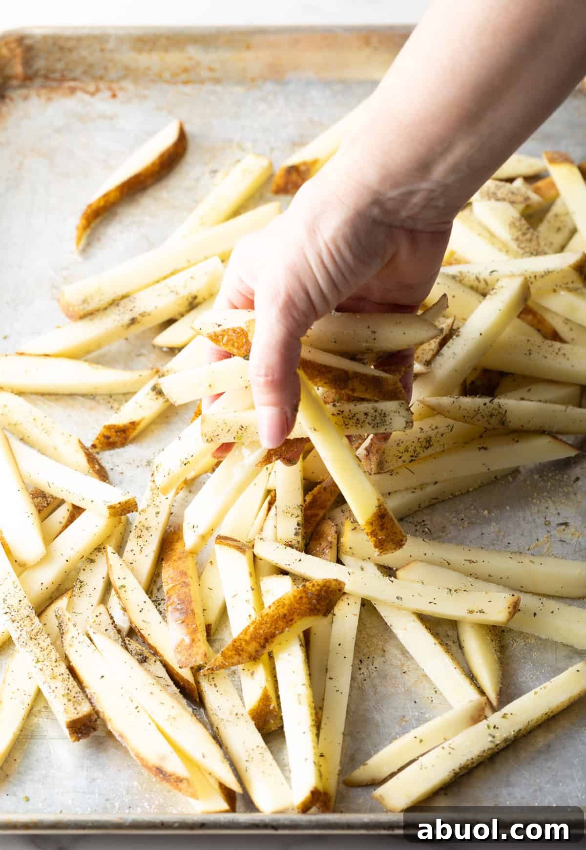 A hand is tossing raw potato slices in oil and seasoning, preparing them for a baked French fries recipe.