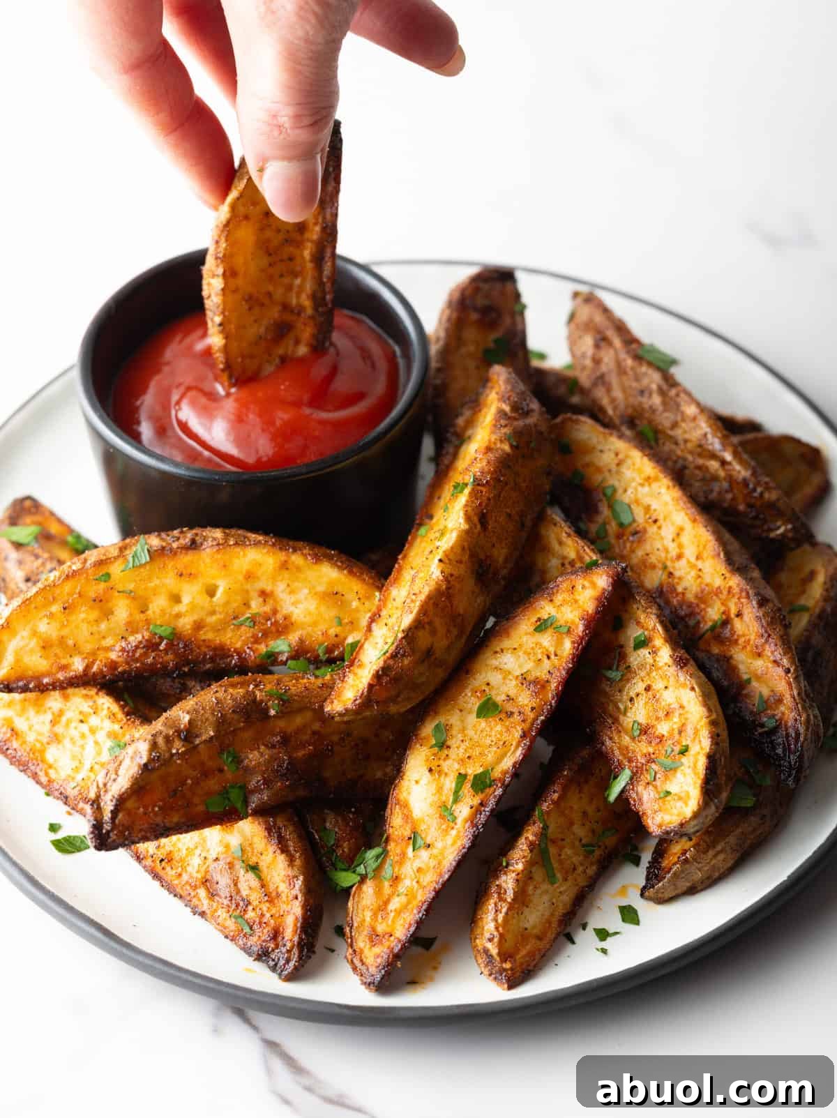 A plate full of oven baked potato wedges with a hand dipping one of the wedges into a ramekin of ketchup.
