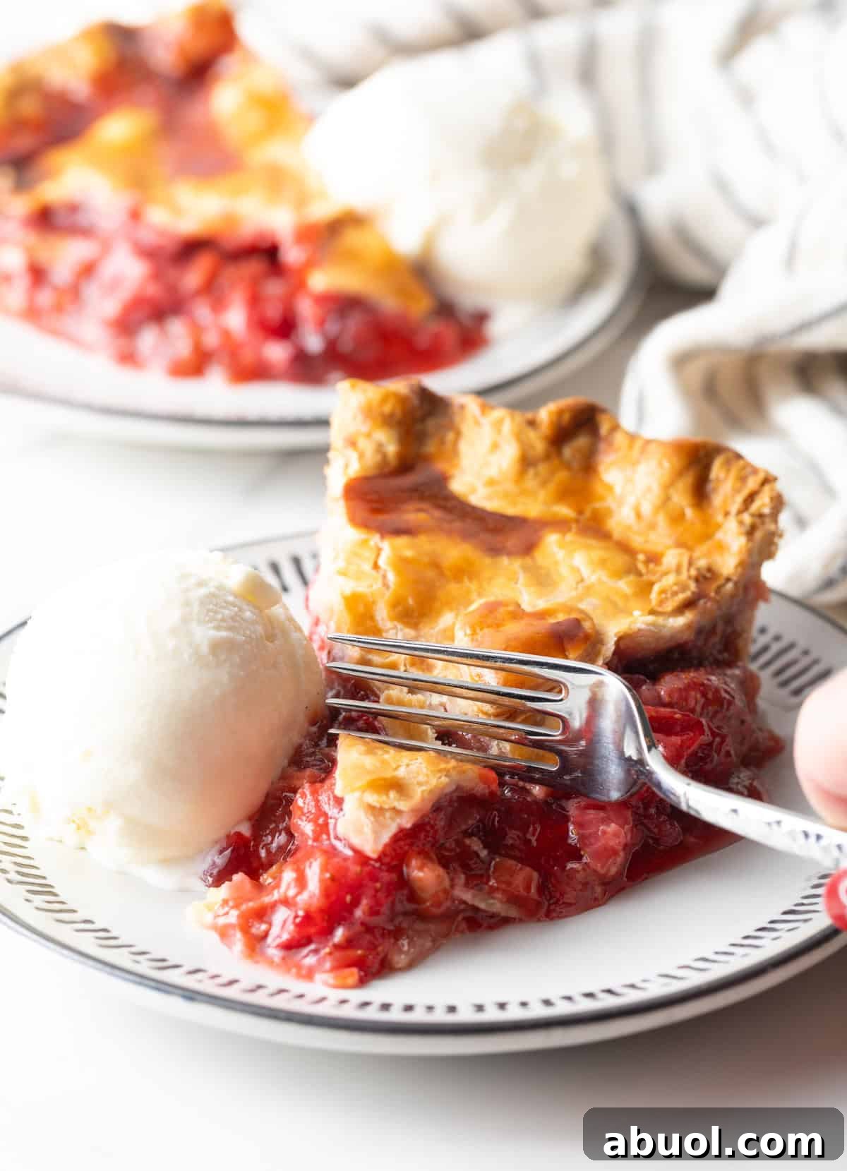 A slice of homemade rhubarb strawberry pie is sitting on a plate with ice cream, and a fork is taking a bite out of the pie.