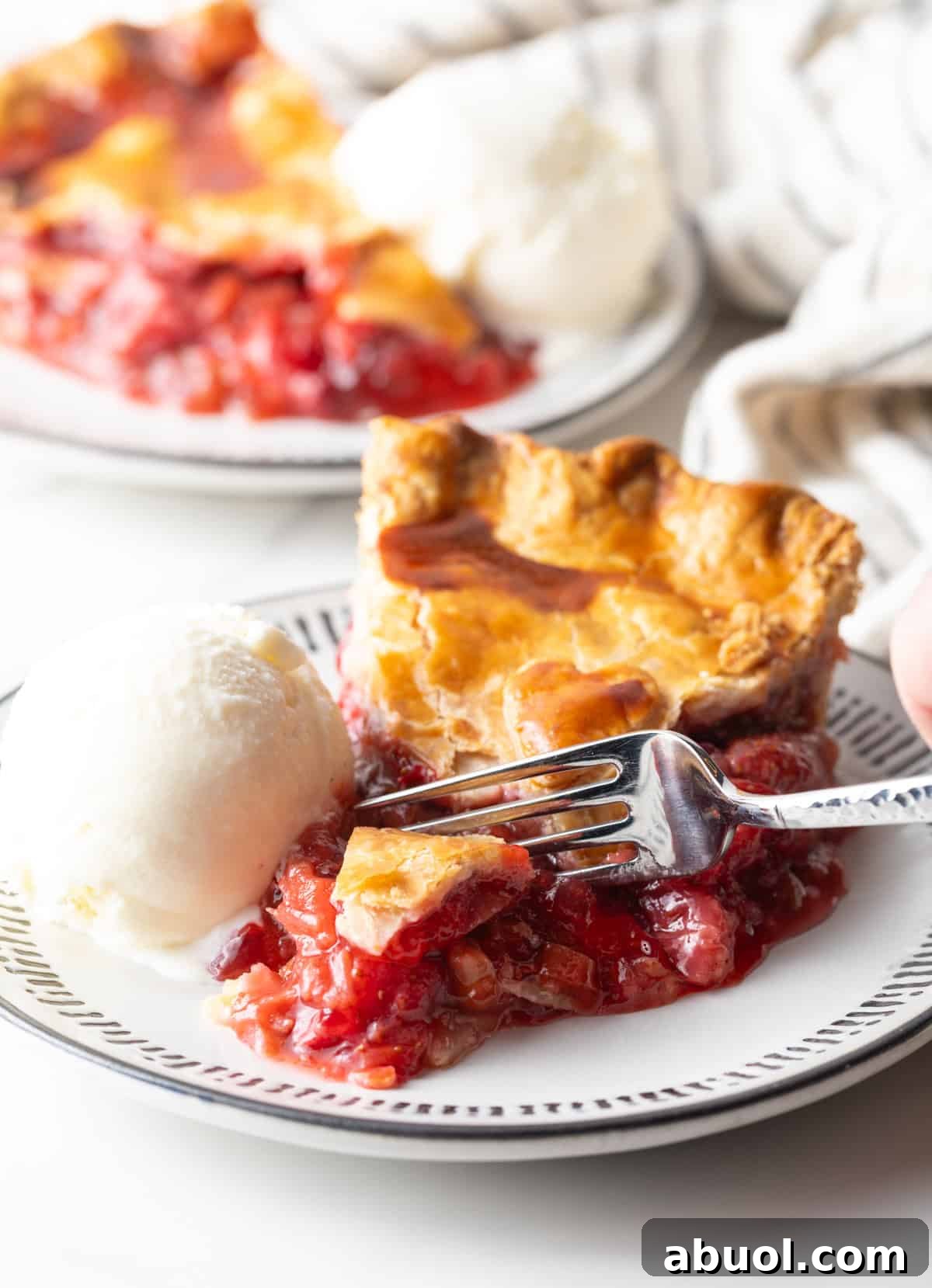 A slice of homemade strawberry rhubarb pie sitting on a plate with ice cream, and a fork is taking a bite out of the pie.
