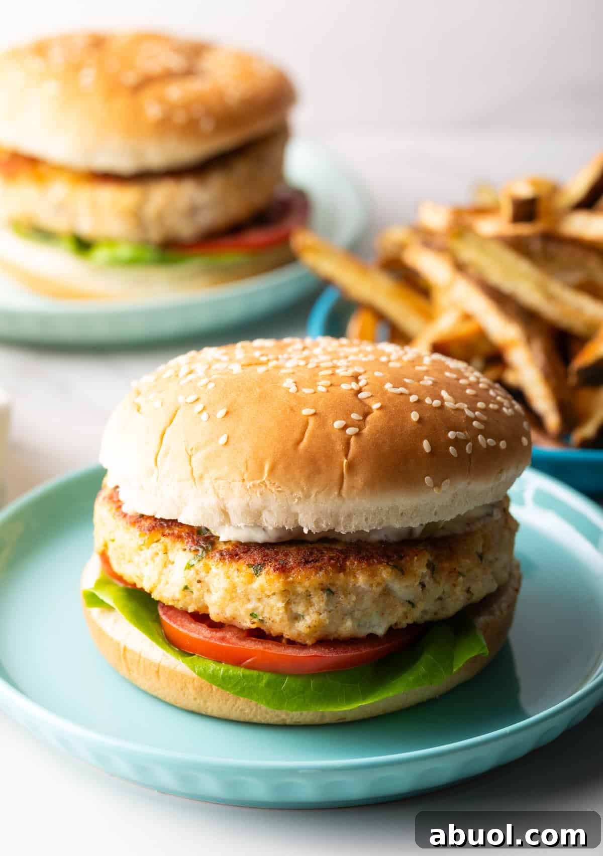 A homemade fish burger with lettuce and tomato on a blue plate, with a plate of fries on the side.