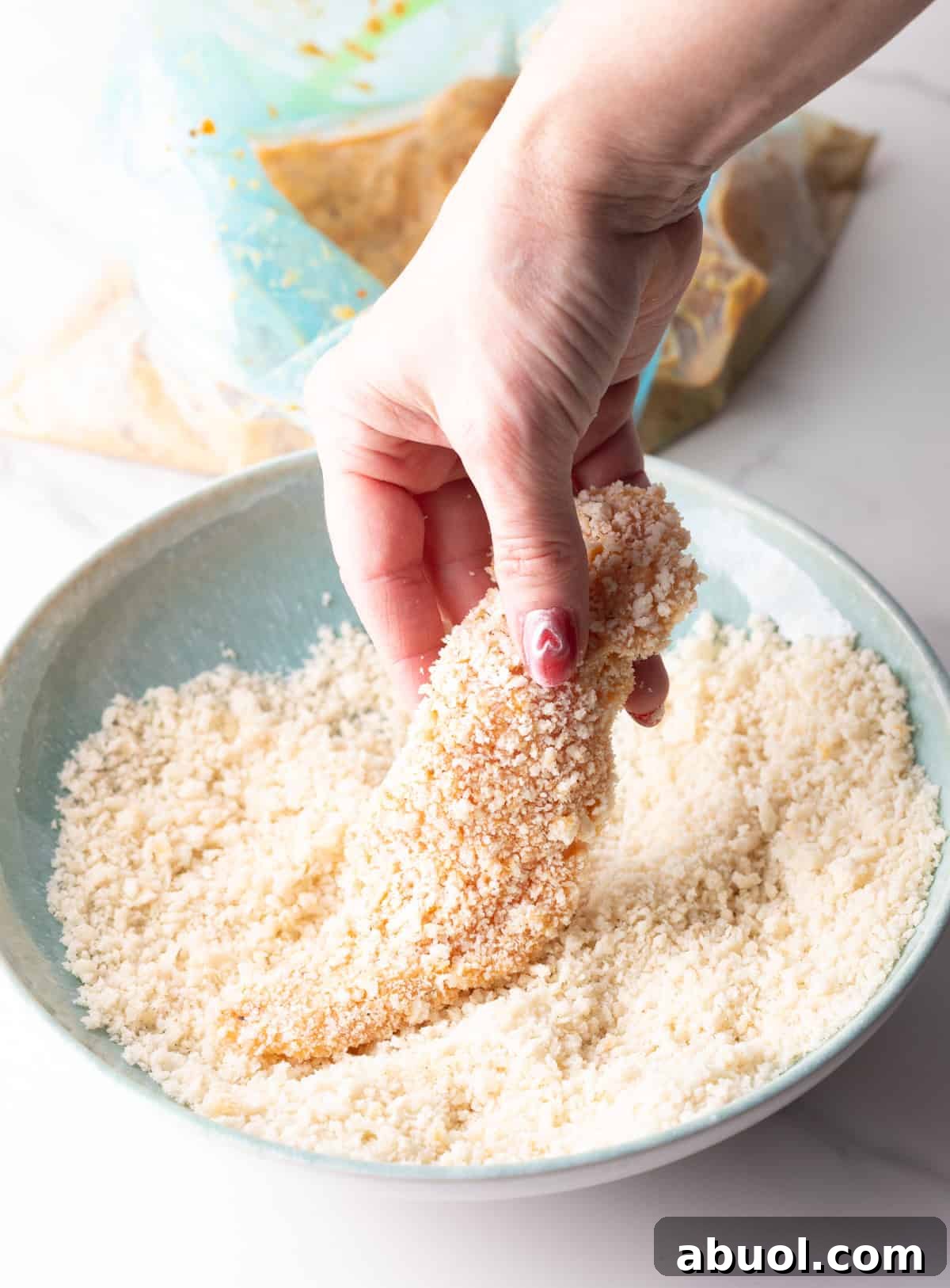 Raw chicken tenders are being coated in panko breadcrumbs for an air fryer chicken tenders recipe.