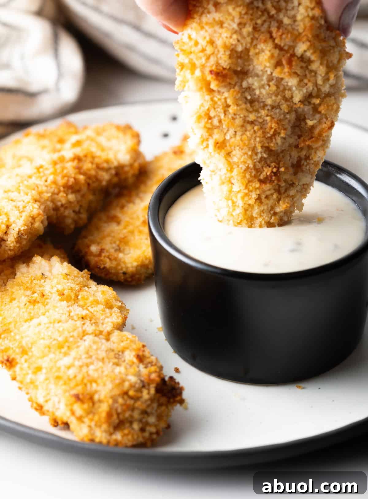A plate of homemade chicken tenders made in the air fryer, with one chicken tender being dipped into a ramekin of white sauce.