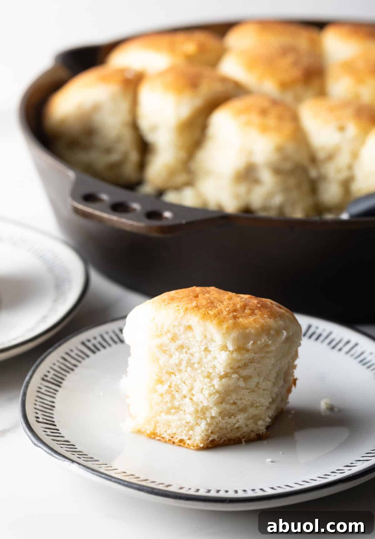 A fluffy homemade angel biscuit is sitting on a white plate, next to a skillet filled with angel biscuits.