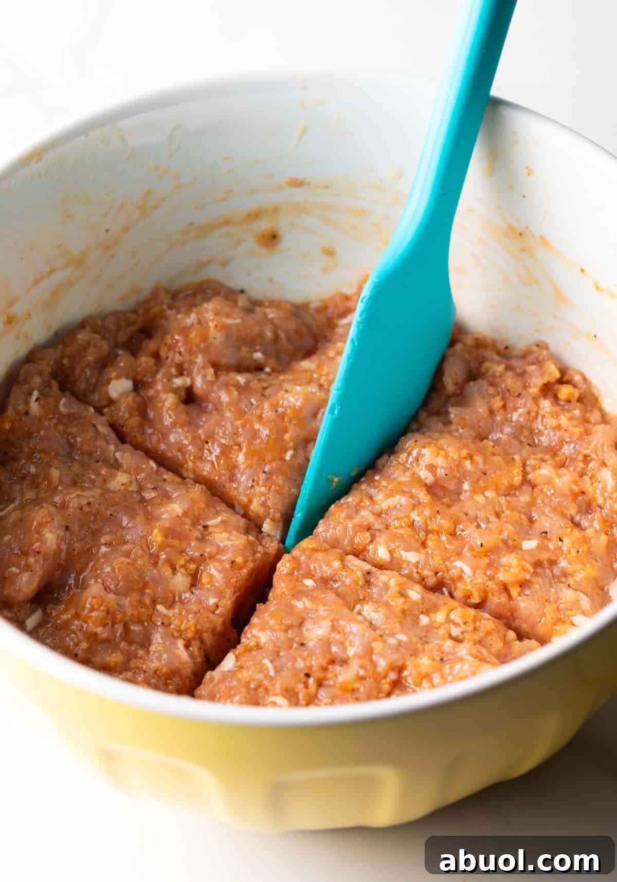 A ground chicken patties mixture in a white bowl, being divided into 4 portions with a blue spatula.