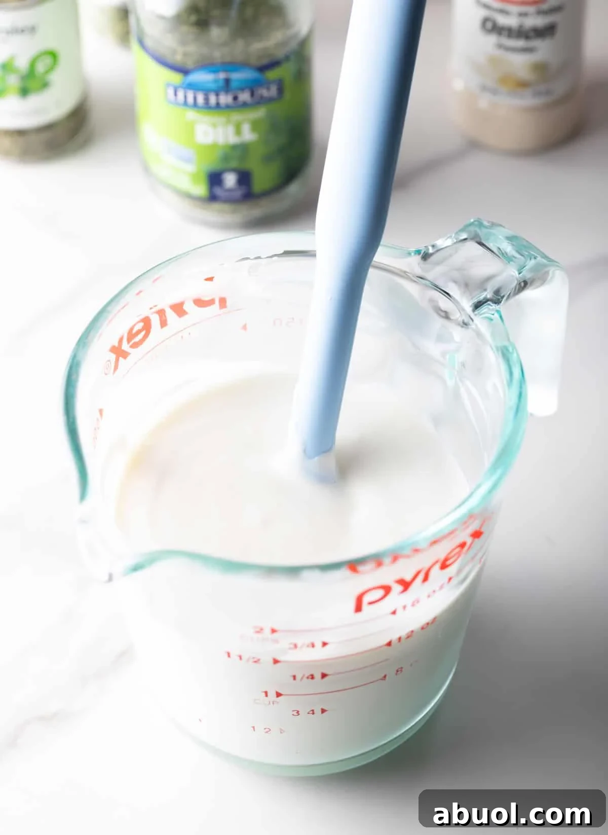 A glass measuring pitcher filled with the creamy base of a buttermilk ranch recipe, being stirred by a spatula.